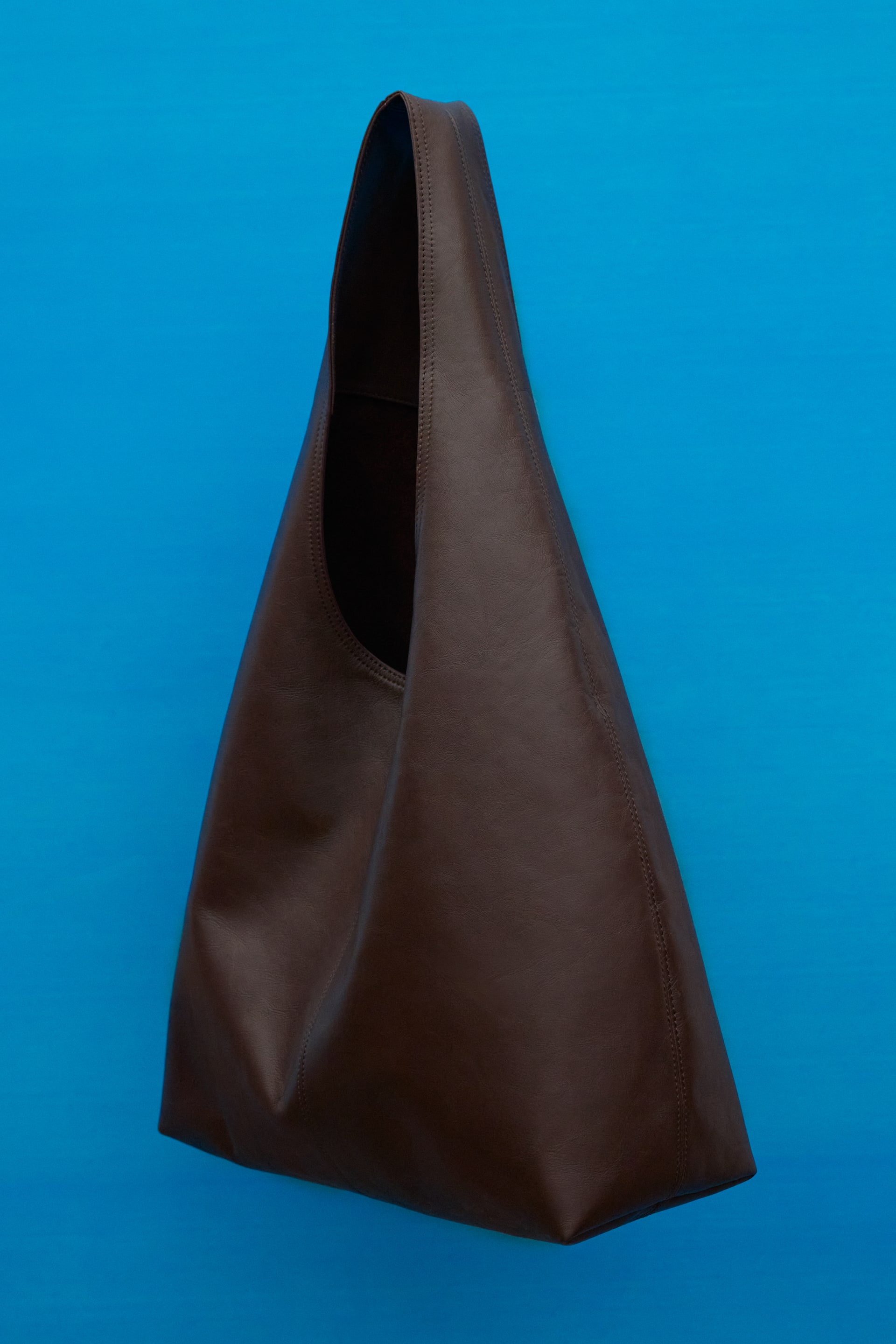 Größeres Bild ansehen: Eine dunkelbraune Leder-Hobo-Tasche hängt und zeigt ihre weiche, unstrukturierte Form. Sie verfügt über einen breiten, integrierten Riemen und sichtbare Nahtstiche.