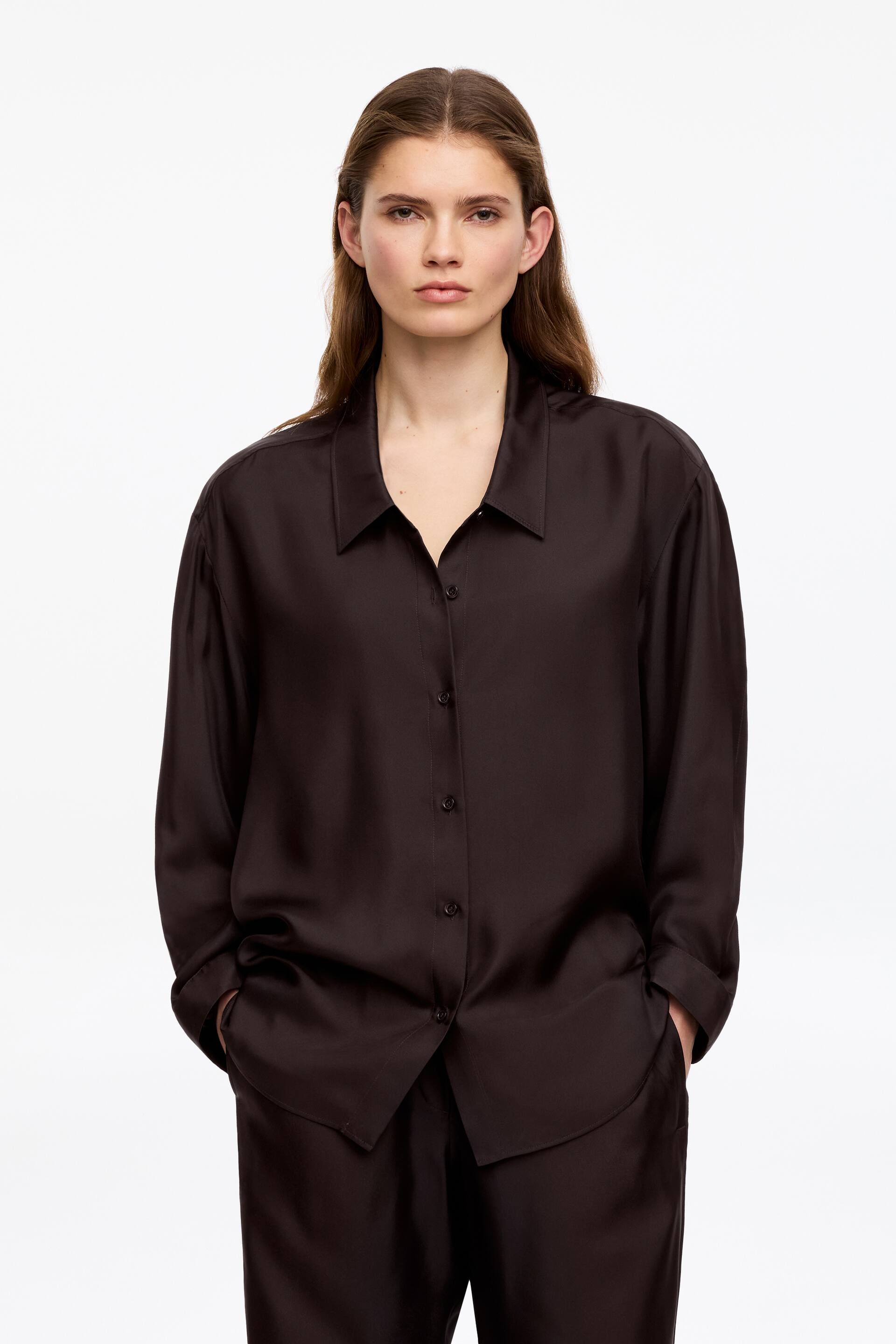Grösseres Bild ansehen: Ein dunkelbraunes Seidenhemd mit tiefem Kragen, Knopfverschluss und abgerundetem Saum wird vom Model getragen. Die Hände des Models ruhen in den Taschen der passenden dunkelbraunen Hose.