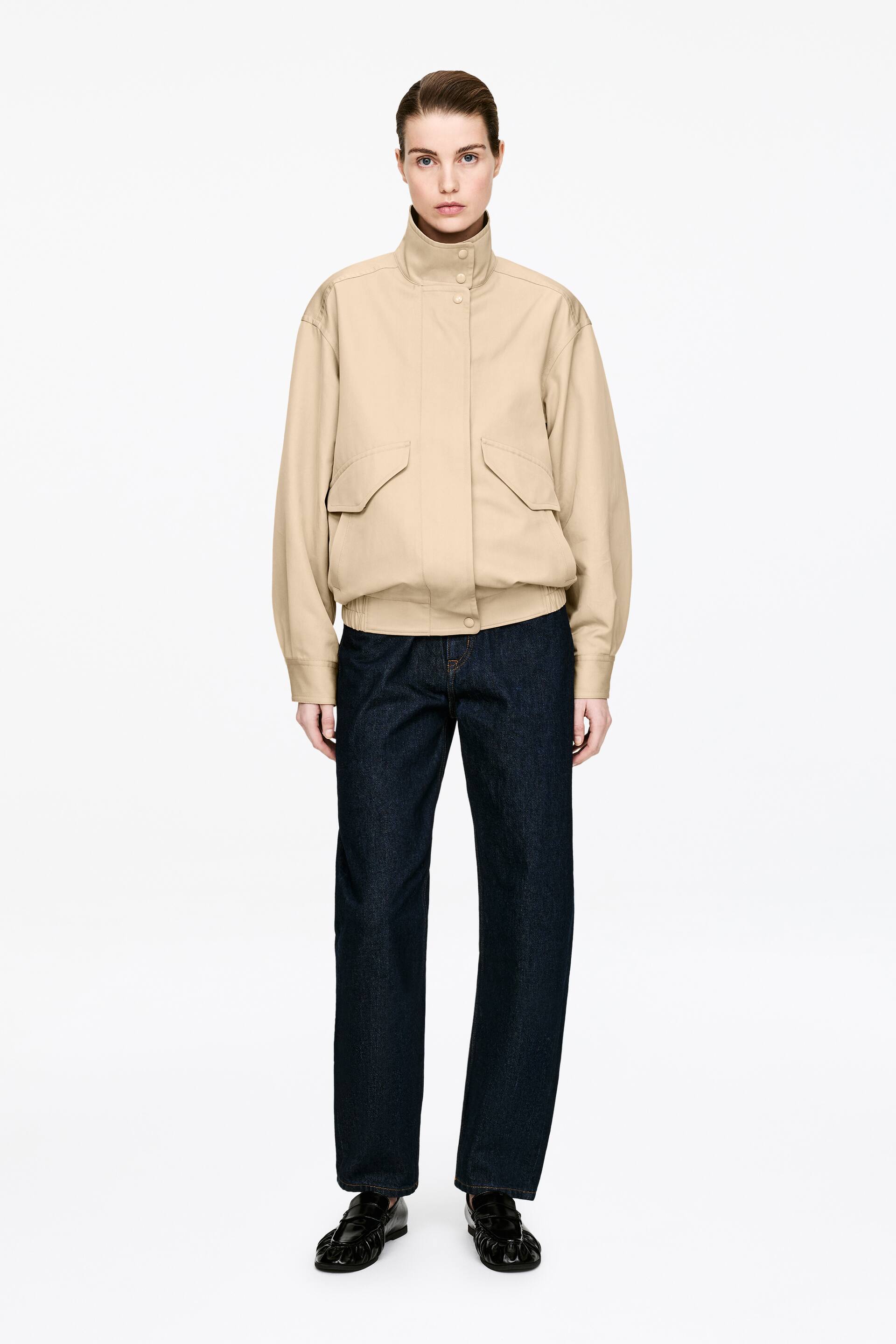 Ver imagen más grande: Una chaqueta bomber beige se presenta de frente, con un cuello alto con botones de presión y una tapeta frontal con broches. Muestra dos grandes bolsillos con solapa en la parte delantera, hombros caídos y puños y dobladillo fruncidos, llevada sobre un top oscuro de cuello alto.