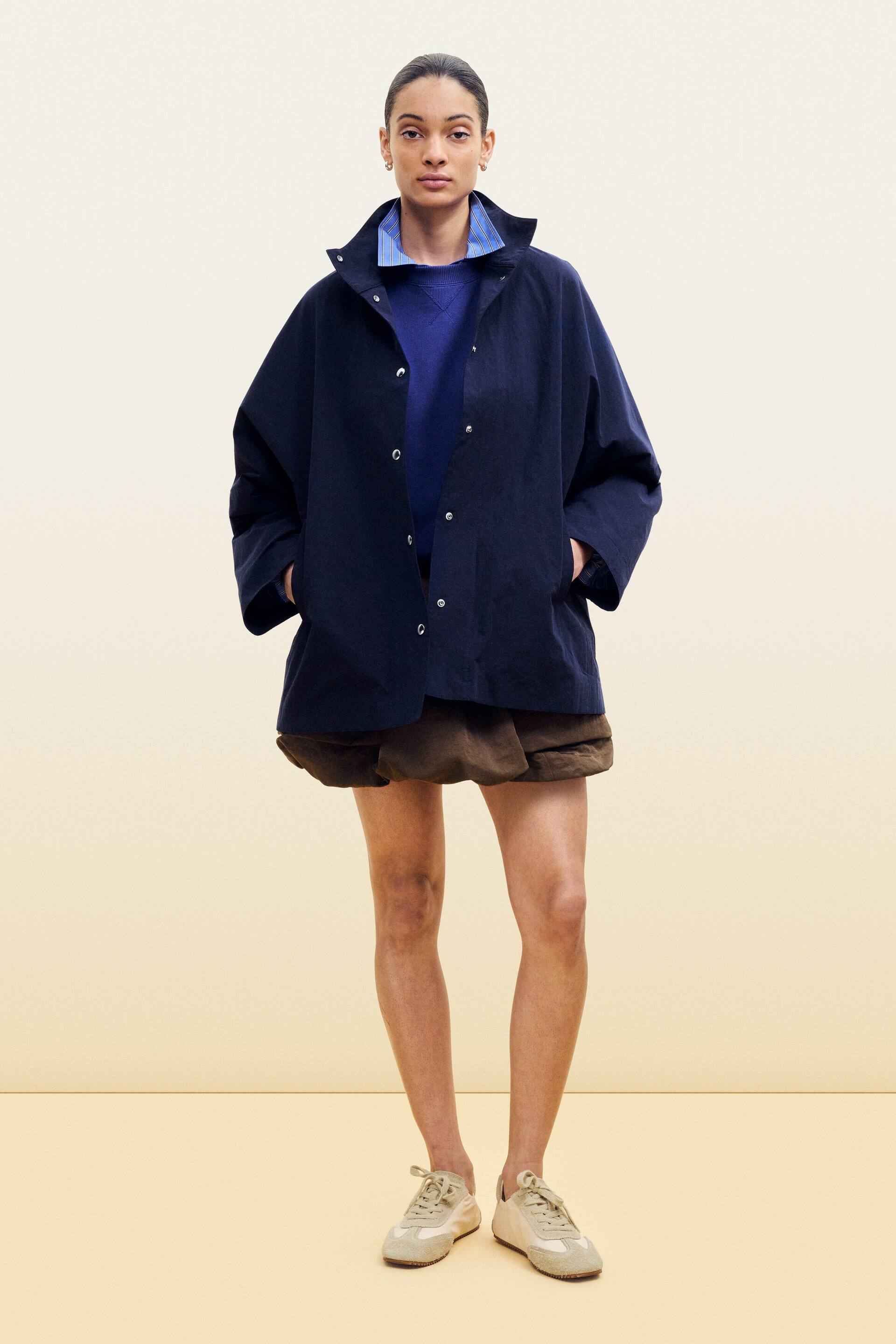 Agrandir l'image: Une veste bleu marine présente une silhouette surdimensionnée avec un col montant et des fermetures à boutons-pression sur le devant, les mains reposant dans ses deux poches avant. Elle est portée ouverte sur un pull ras du cou bleu, avec une chemise à col rayé bleu clair visible sous le col de la veste.