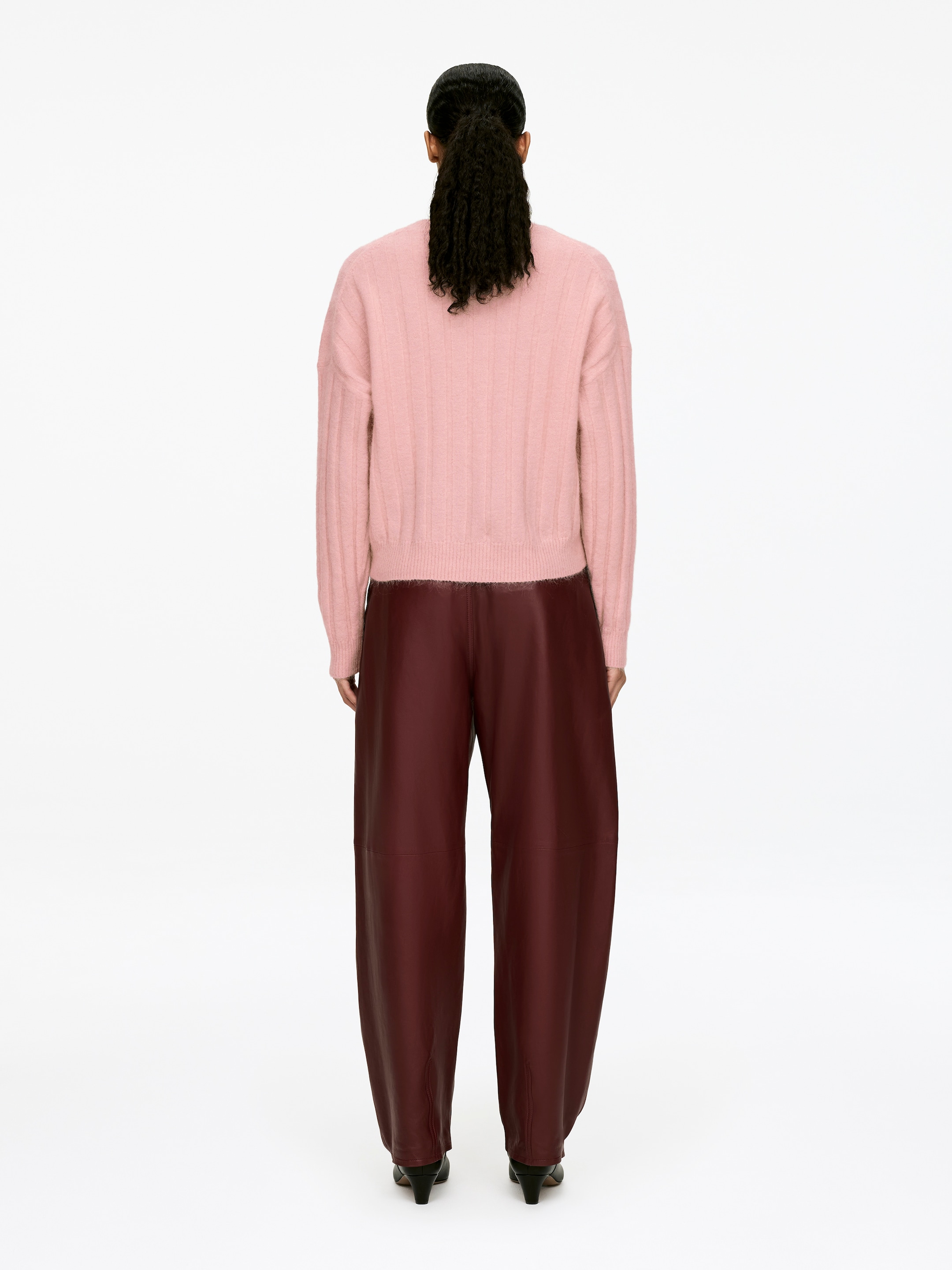 Grösseres Bild ansehen: Ein pinkfarbener, breiter Rippstrickpullover mit vertikaler Rippung und entspanntem Schnitt wird vom Model getragen. Der Saum des langärmeligen Pullovers liegt über dunkelburgunderfarbenen Hosen.