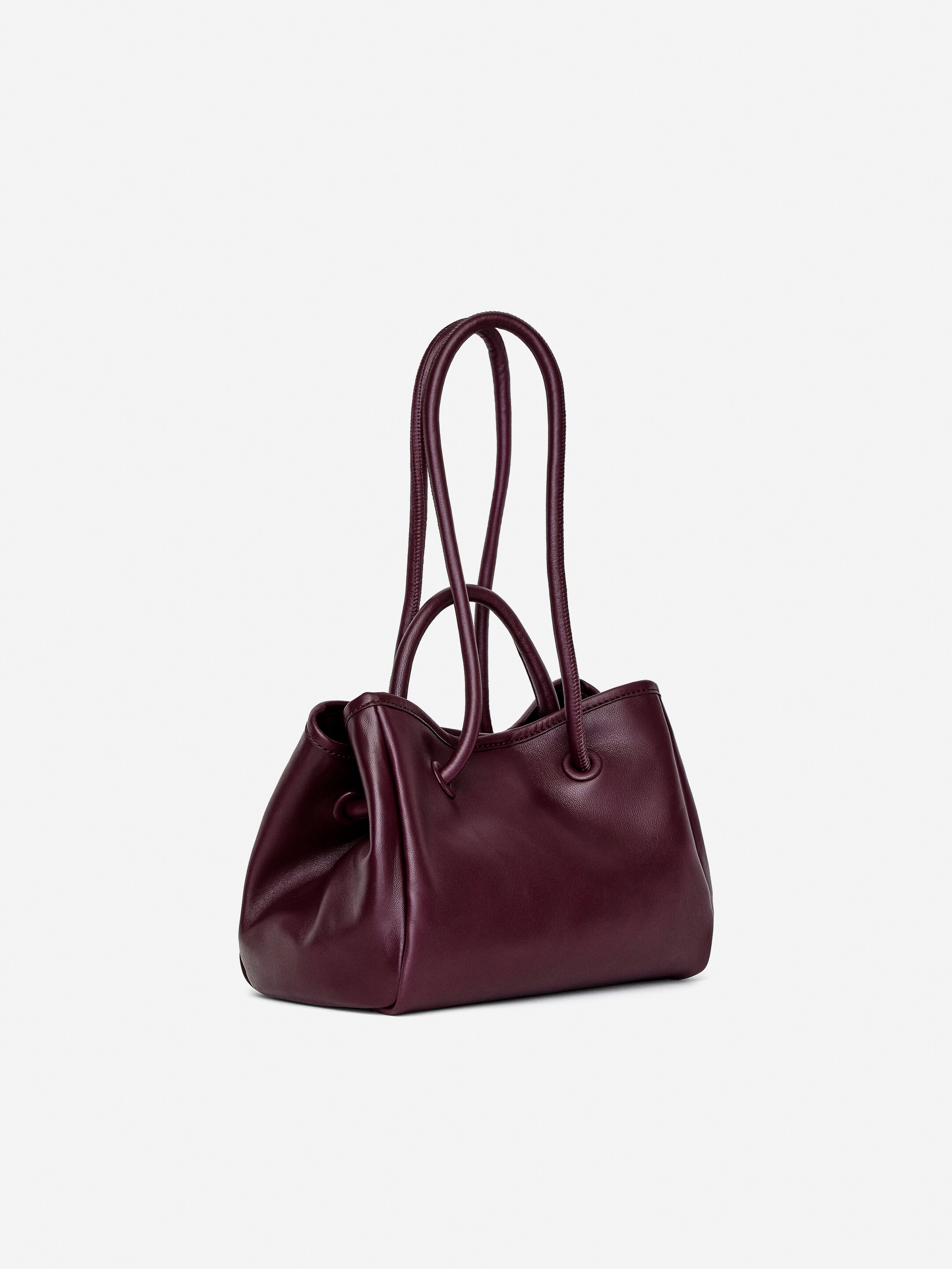 Visualizza immagine più grande: Una borsa a tracolla in pelle bordeaux scuro è rivolta a sinistra, con manici superiori e tracolle tubolari che scorrono attraverso occhielli.