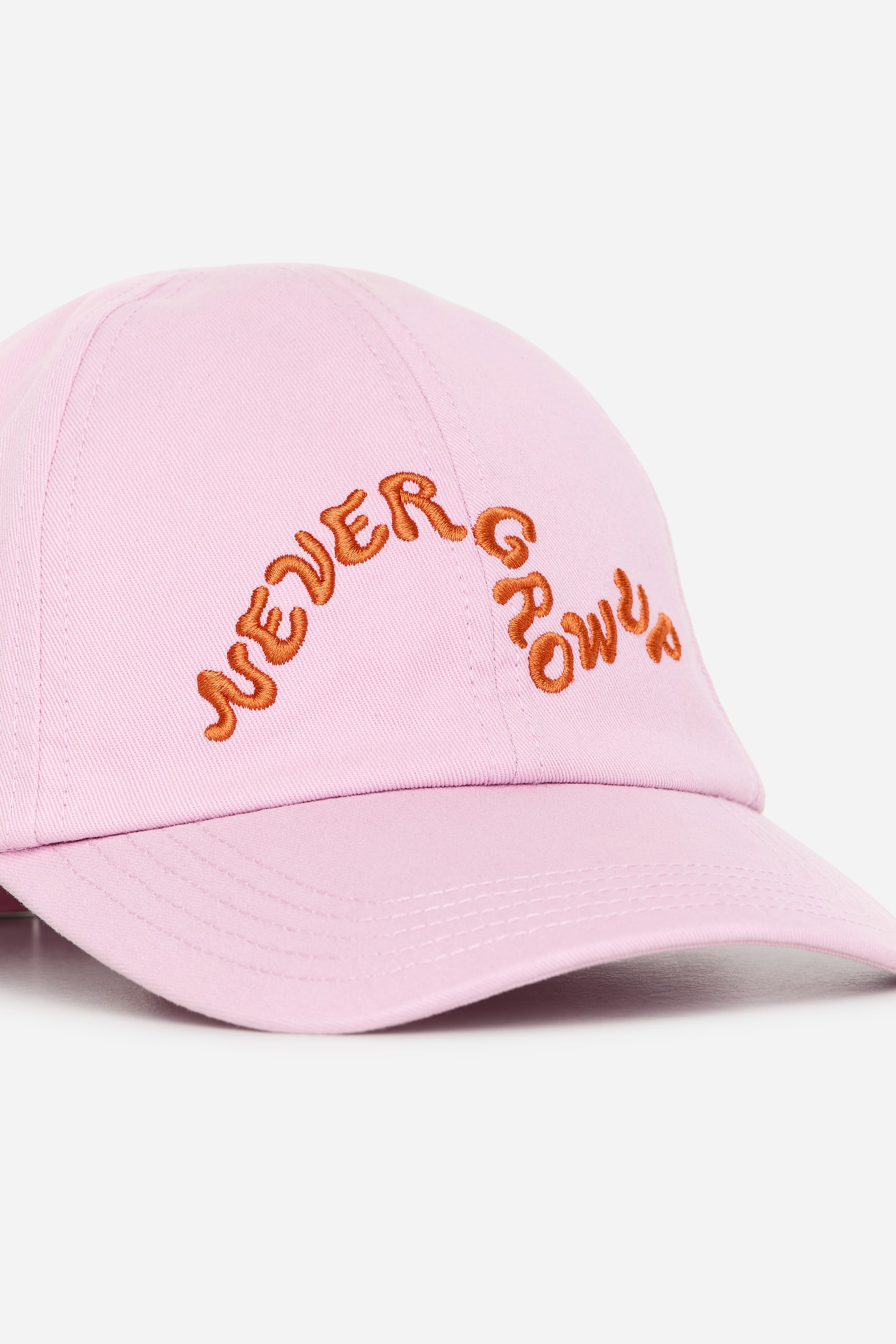 Grösseres Bild ansehen: Eine hellrosa Baseball-Cap zeigt ihre Vorderansicht und präsentiert orangefarbene Stickschrift mit dem Text "NEVER GROW UP", der sich über das Hauptpanel wölbt. Die Cap verfügt außerdem über einen gebogenen Schirm mit sichtbaren Nahtlinien.
