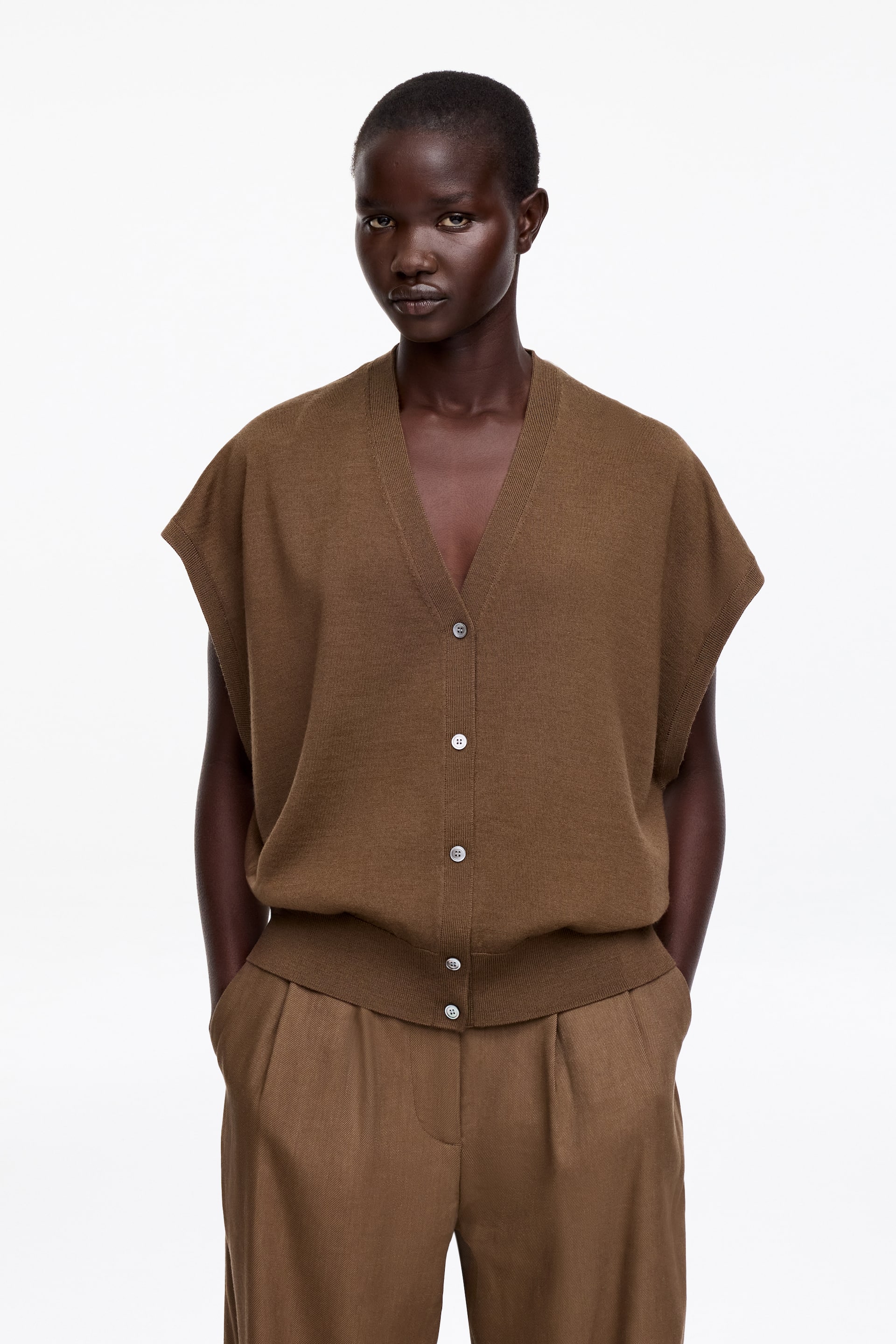 Visa större bild: En brun stickad koftväst med djup V-ringning och förlängda axlar. Den är designad med en knappstängning framtill, säkrad med fem ljusa knappar, och definieras av en ribbad fåll, som visar upp en avslappnad passform.