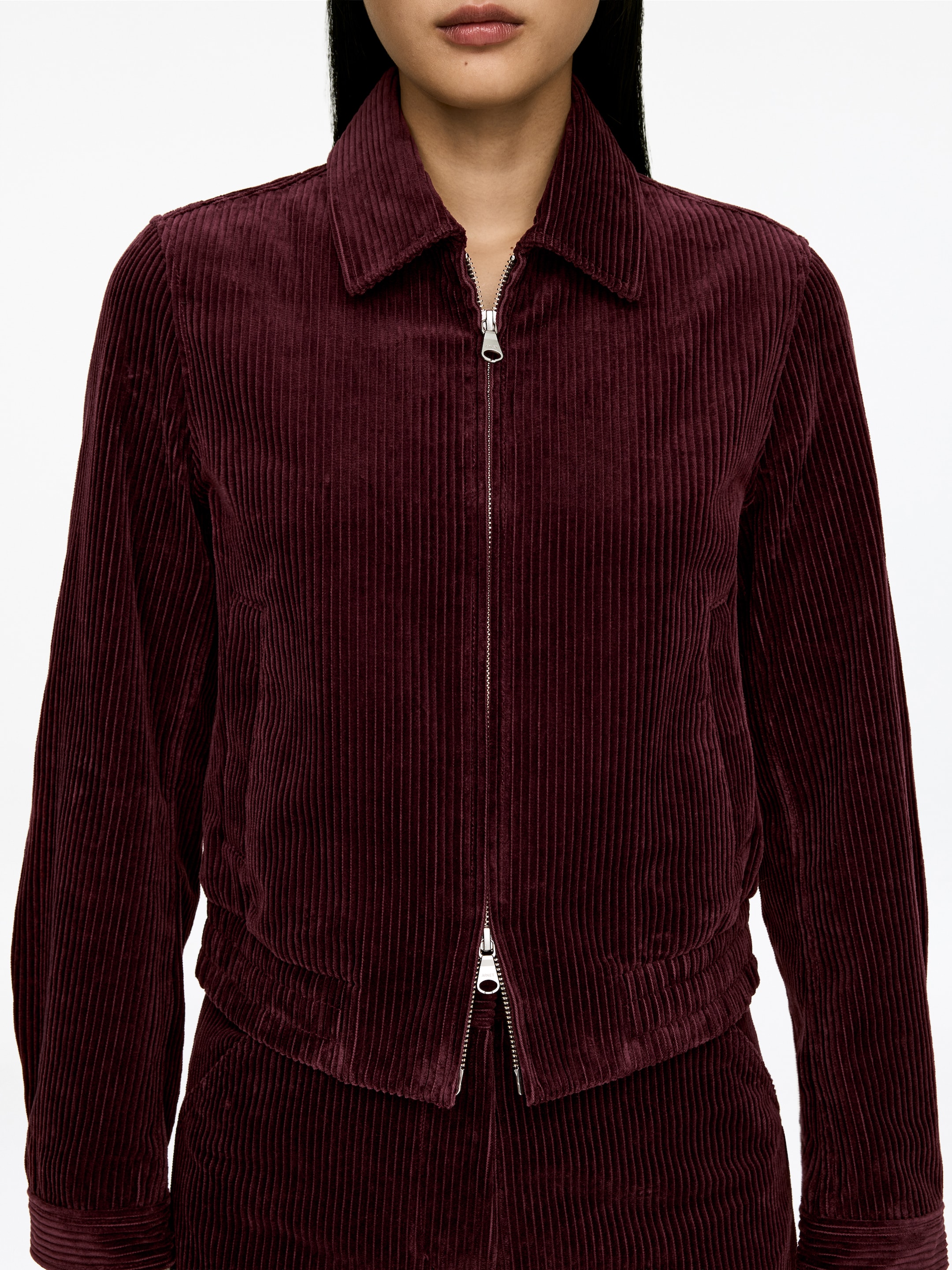 Größeres Bild ansehen: Eine Cordjacke mit breitem Rippenmuster in Burgund wird von vorne präsentiert. Sie verfügt über einen Reißverschluss mit silberfarbenem Zipper, einen spitzen Kragen, lange Ärmel und einen gerafften Saum.