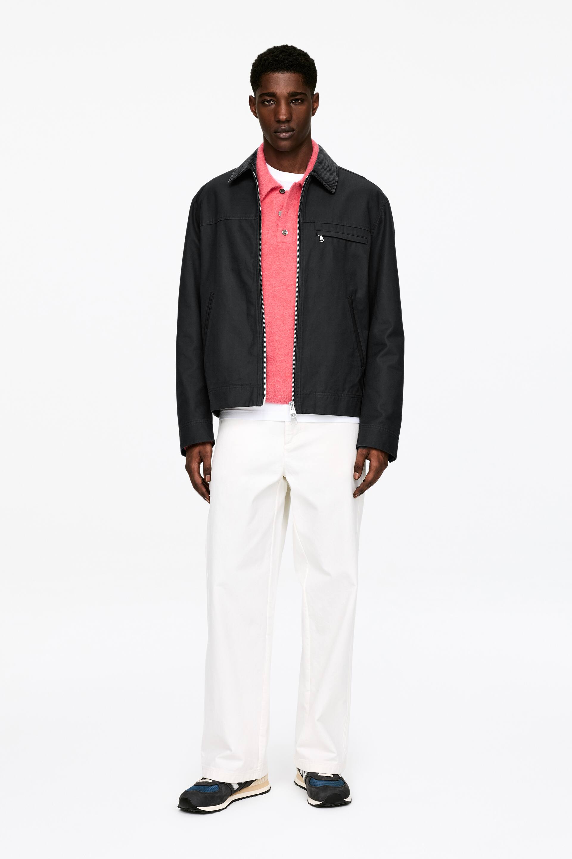 Agrandir l'image: Une veste noire de style workwear est présentée, face à l'avant. Elle est dotée d'une fermeture éclair complète à l'avant, d'un col évasé foncé contrastant et de deux poches poitrine zippées. La veste est portée ouverte, révélant un polo rose et un t-shirt blanc en dessous.