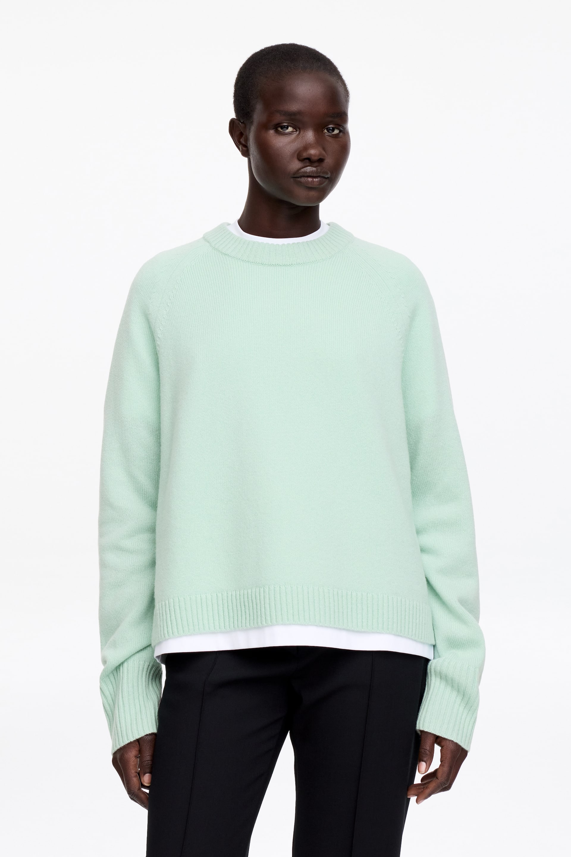 Ver imagen más grande: Un jersey de punto verde claro presenta un cuello redondo acanalado, mangas raglán, puños acanalados y un dobladillo acanalado. Está superpuesto sobre una camiseta blanca, con el dobladillo de la camiseta visible debajo del jersey.