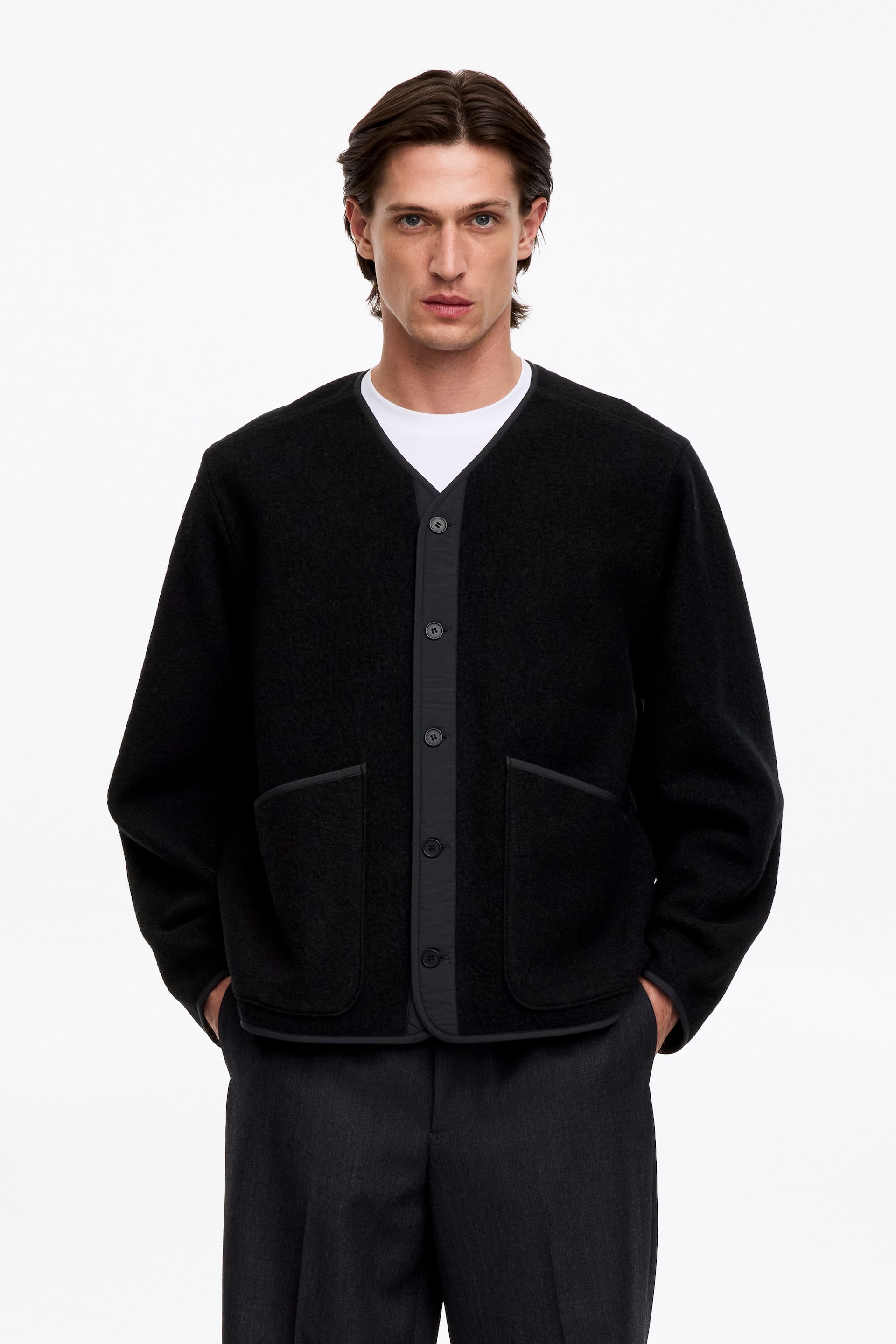 Grotere afbeelding bekijken: Een zwart cardigan met textuur, een V-hals, een contrasterende knoopsluiting aan de voorkant en twee opgestikte zakken met bies wordt door het model gedragen over een wit T-shirt.