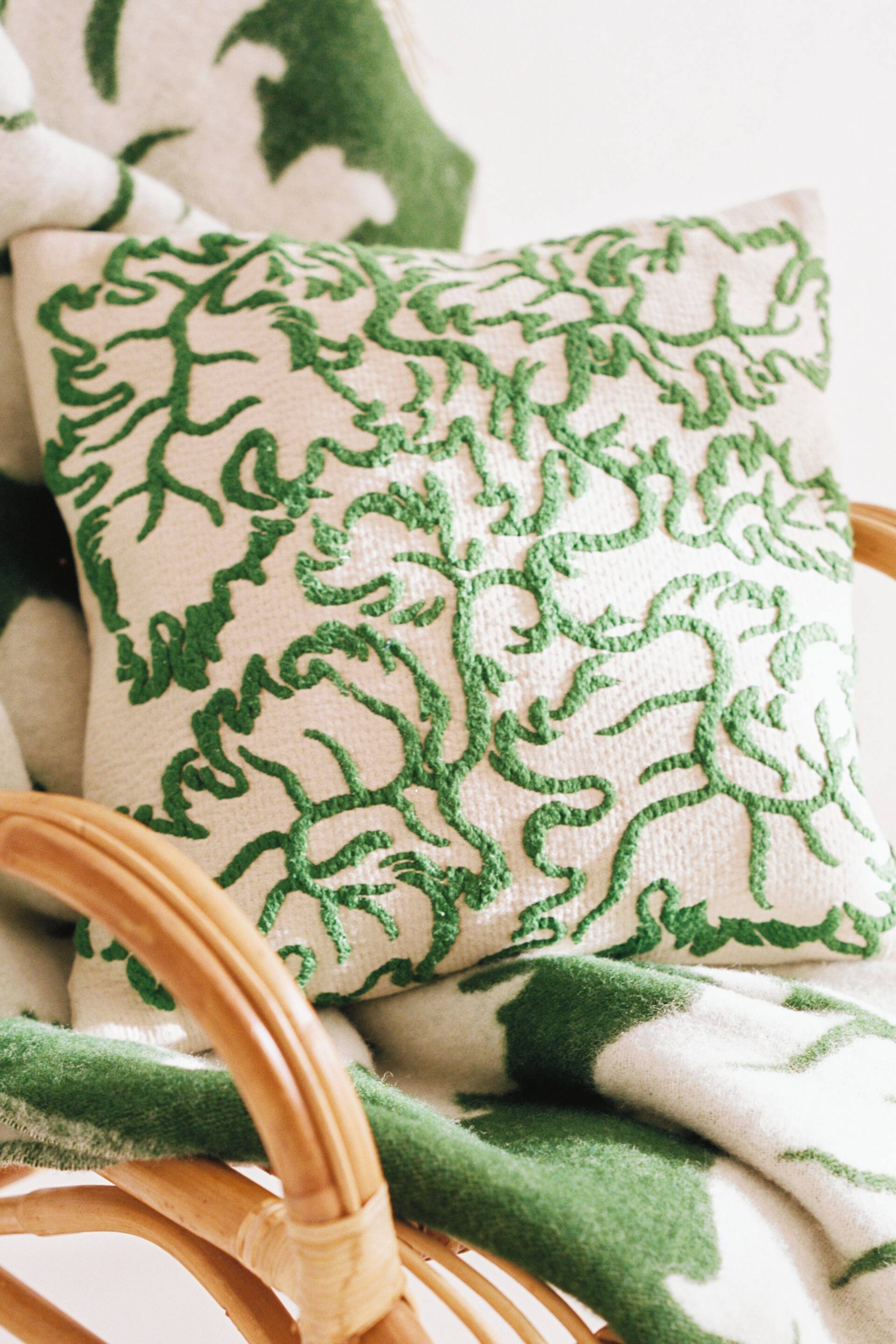 Vis større bilde: En kvadratisk kremfarget pyntepute med et tett, hevet grønt broderimønster med abstrakte, organiske former. Den er plassert oppreist, hvilende på et matchende krem- og grønnmønstret teppe.