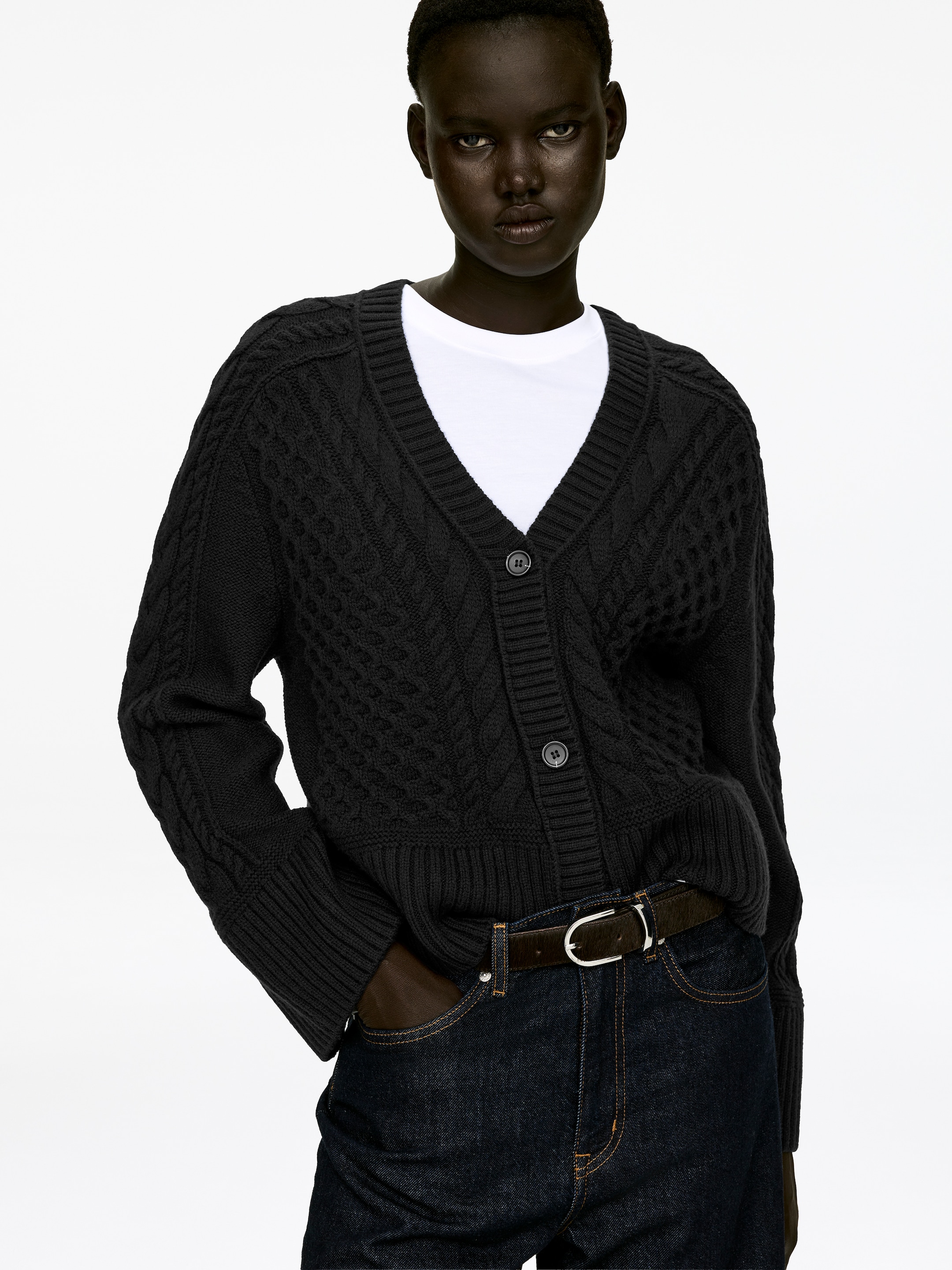Grösseres Bild ansehen: Eine schwarze Zopfstrickjacke mit V-Ausschnitt, Drei-Knopf-Verschluss und gerippten Bündchen wird vom Model getragen. Die Strickjacke ist über ein weißes T-Shirt geschichtet, und das Model steckt eine Hand in eine Jeans-Tasche.