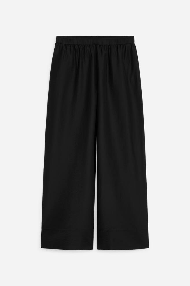 Streamlined black trousers, crafted from a lyocell blend, are showcased from the front. They are designed with an elasticated waist, side seam pockets, and a single welt back pocket with button fastening, featuring a wide-leg and cropped silhouette. - 1
