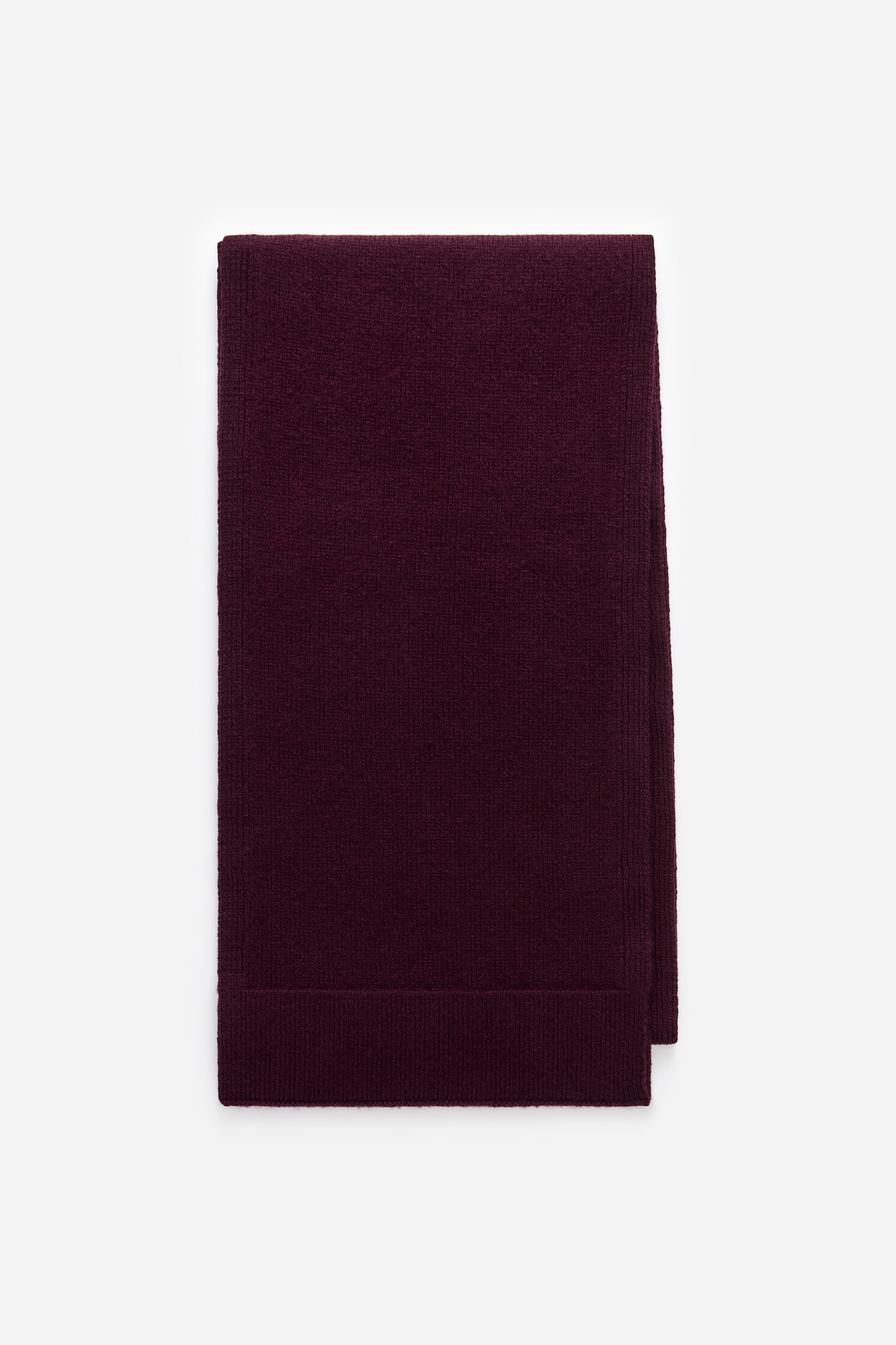 Größeres Bild ansehen: Nativa Wollschal. Die Vorderseite dieses dunkelvioletten Schals ist zu einem kompakten Rechteck gefaltet. Er zeigt eine glatte, feine Strickstruktur mit einem markanten gerippten Band an seinem sichtbaren unteren Rand.