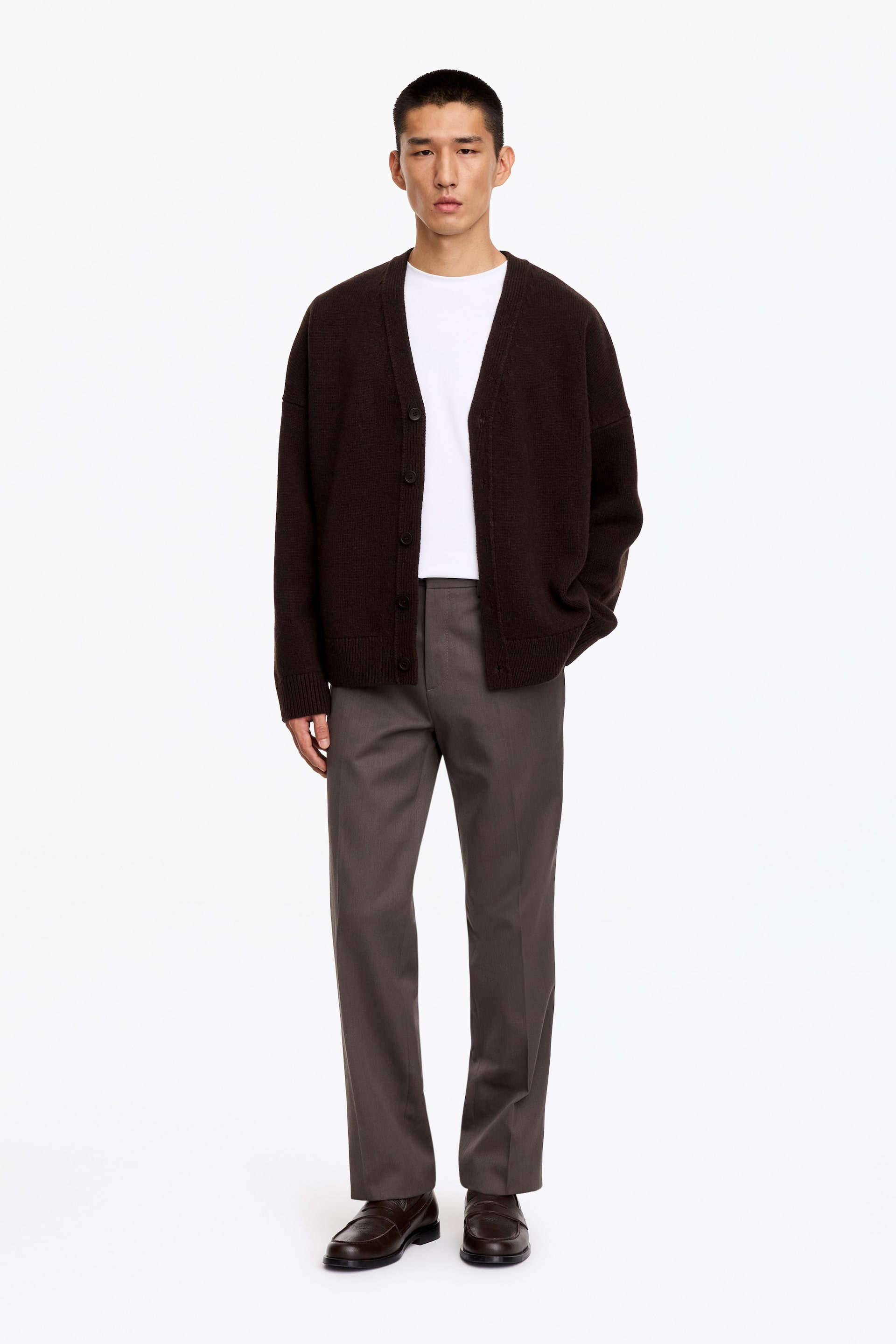 Agrandir l'image: Le mannequin porte un pantalon en mélange laine-coton dans une teinte gris foncé, avec une coupe droite et des plis marqués au centre des jambes. Le pantalon est associé à un t-shirt blanc rentré, un cardigan marron foncé porté ouvert et des mocassins en cuir marron foncé.