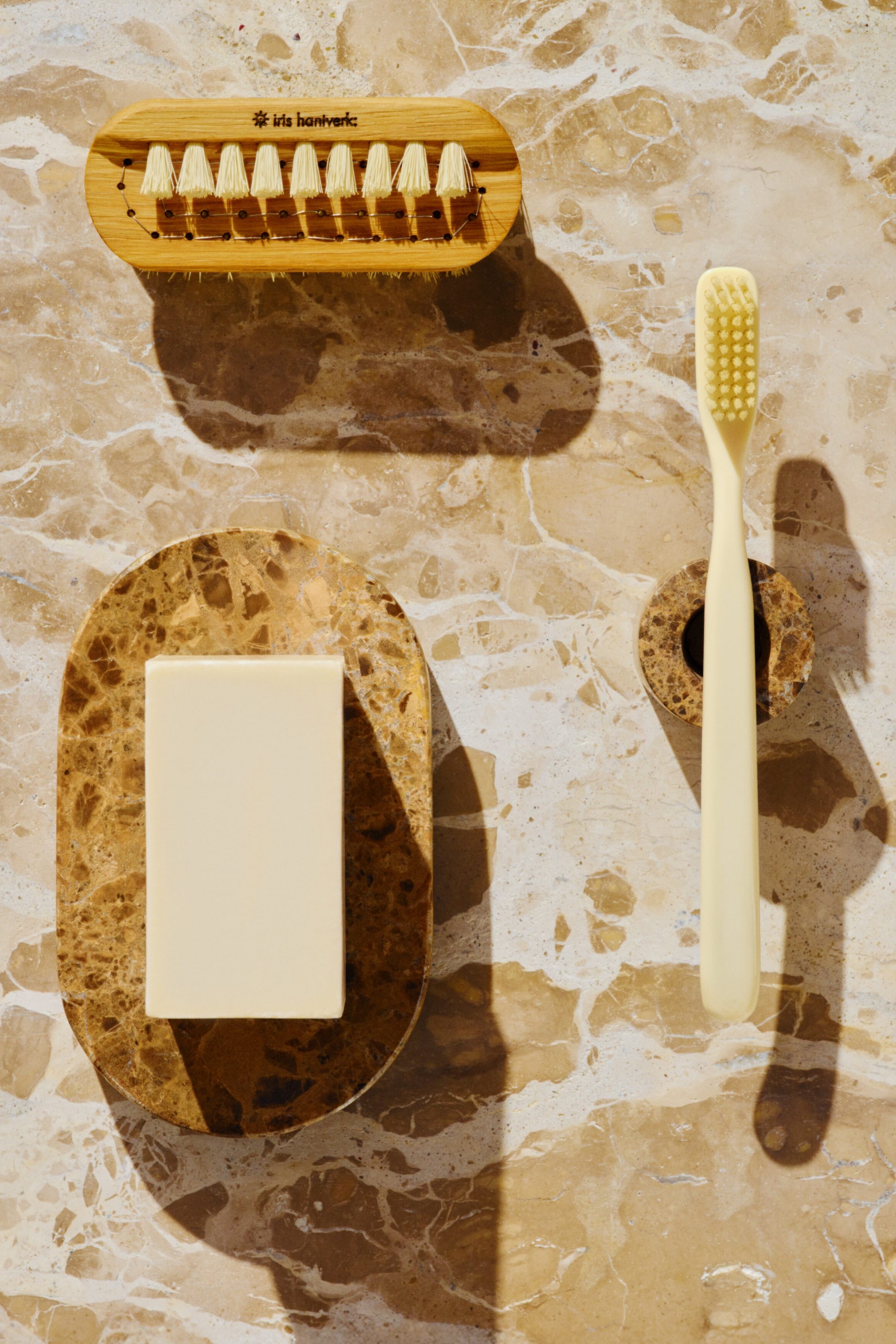 Grösseres Bild ansehen: Eine Holznagelbürste mit cremefarbenen Borsten zeigt den Schriftzug „iris hantverk“. Ein rechteckiges, hellcremefarbenes Seifenstück ruht auf einer ovalen, braunen Marmorseifenschale, und eine cremefarbene Zahnbürste steht aufrecht in einem zylindrischen, braunen Marmor-Zahnbürstenhalter.