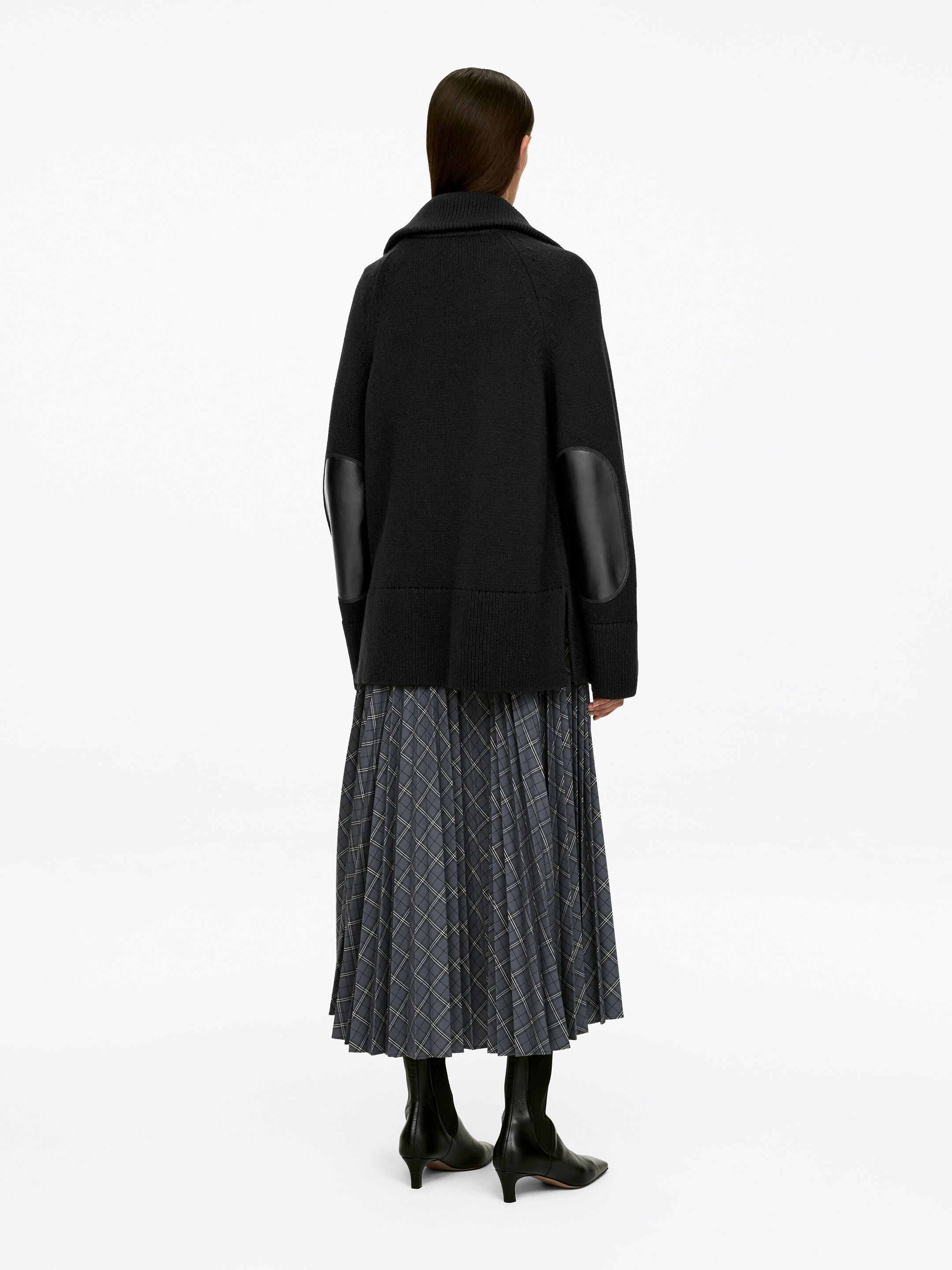 Visualizza immagine più grande: Un maglione nero a maglia con un taglio comodo è indossato dalla modella, visto di schiena. Presenta maniche raglan, un ampio collo a coste e toppe in pelle nera sui gomiti. Il maglione è sovrapposto a una gonna a pieghe a quadretti.