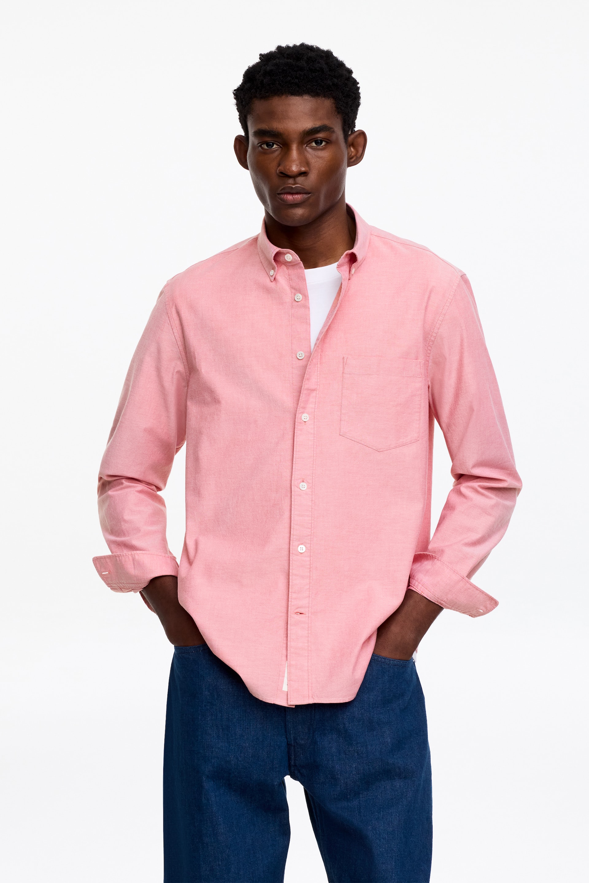 Größeres Bild ansehen: Ein rosa Hemd mit Knopfleiste verfügt über einen Button-Down-Kragen, weiße Knöpfe an der vorderen Knopfleiste und eine einzelne Brusttasche. Die langen Ärmel sind an den Manschetten hochgekrempelt, und es wird über einem weißen T-Shirt getragen, das teilweise in dunkelblaue Jeans gesteckt ist.