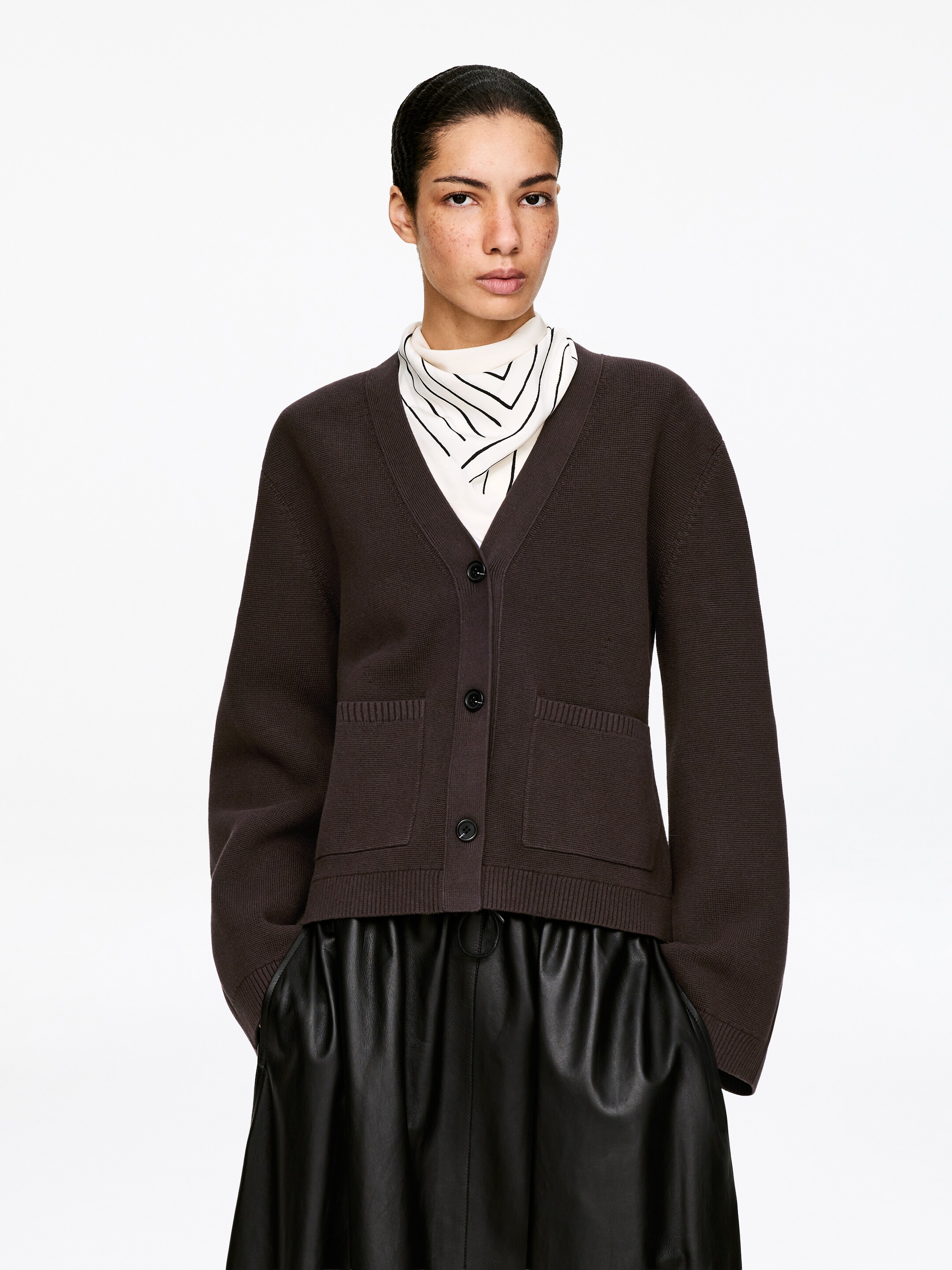 Visa större bild: En mörkbrun kofta visas framifrån, med en V-ringning, mörka runda knappar längs framsidan och två påsydda fickor. Den har ribbade muddar och fåll, överlagrad på en krämfärgad topp med ett svart geometriskt mönster, och kombinerad med svarta veckade läderbyxor.