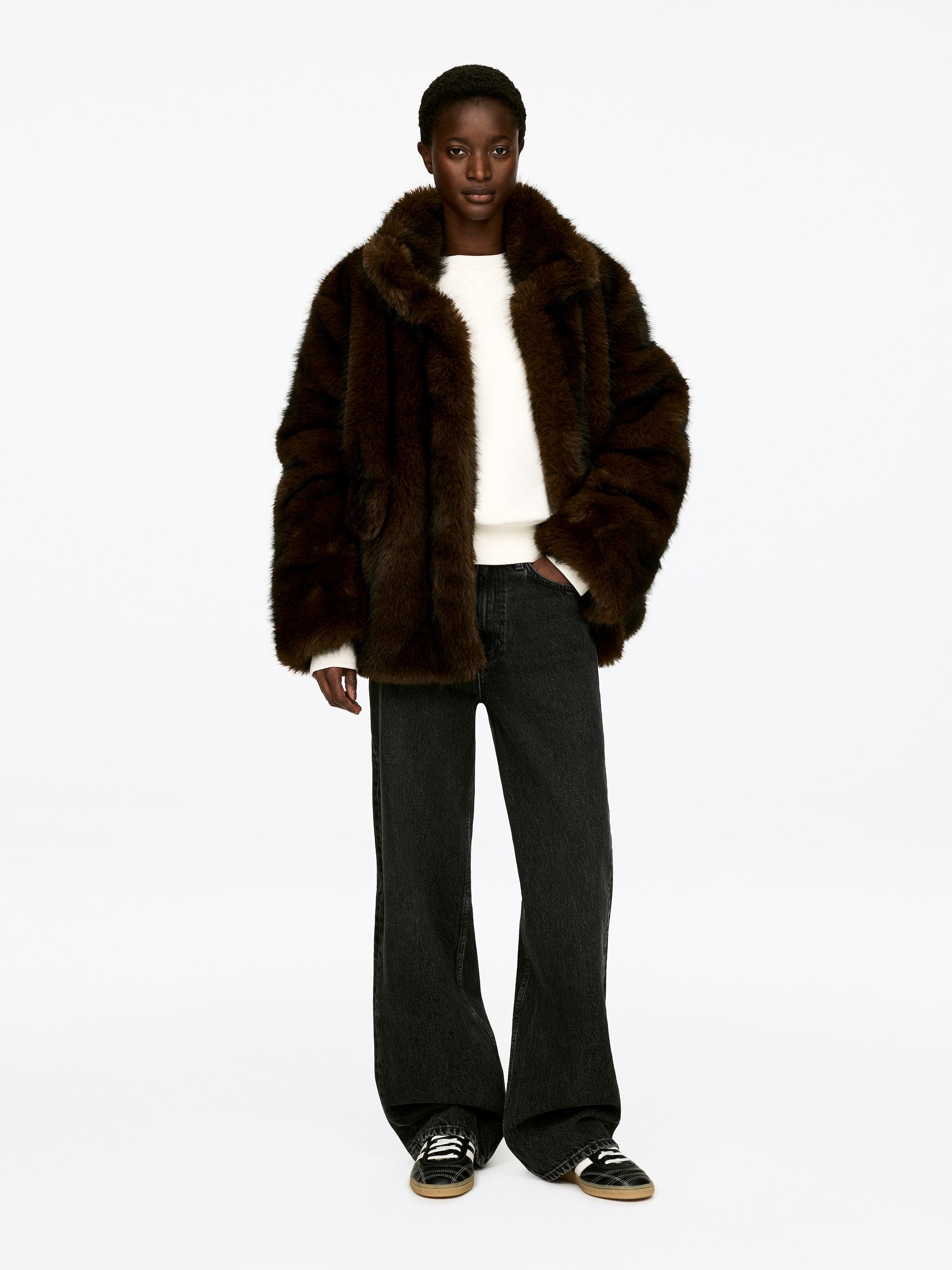 Agrandir l'image: Un manteau en fausse fourrure marron foncé est tourné vers l'avant, avec un col large et une coupe ample et décontractée. Il est porté ouvert, révélant deux poches latérales et un pull blanc en dessous.