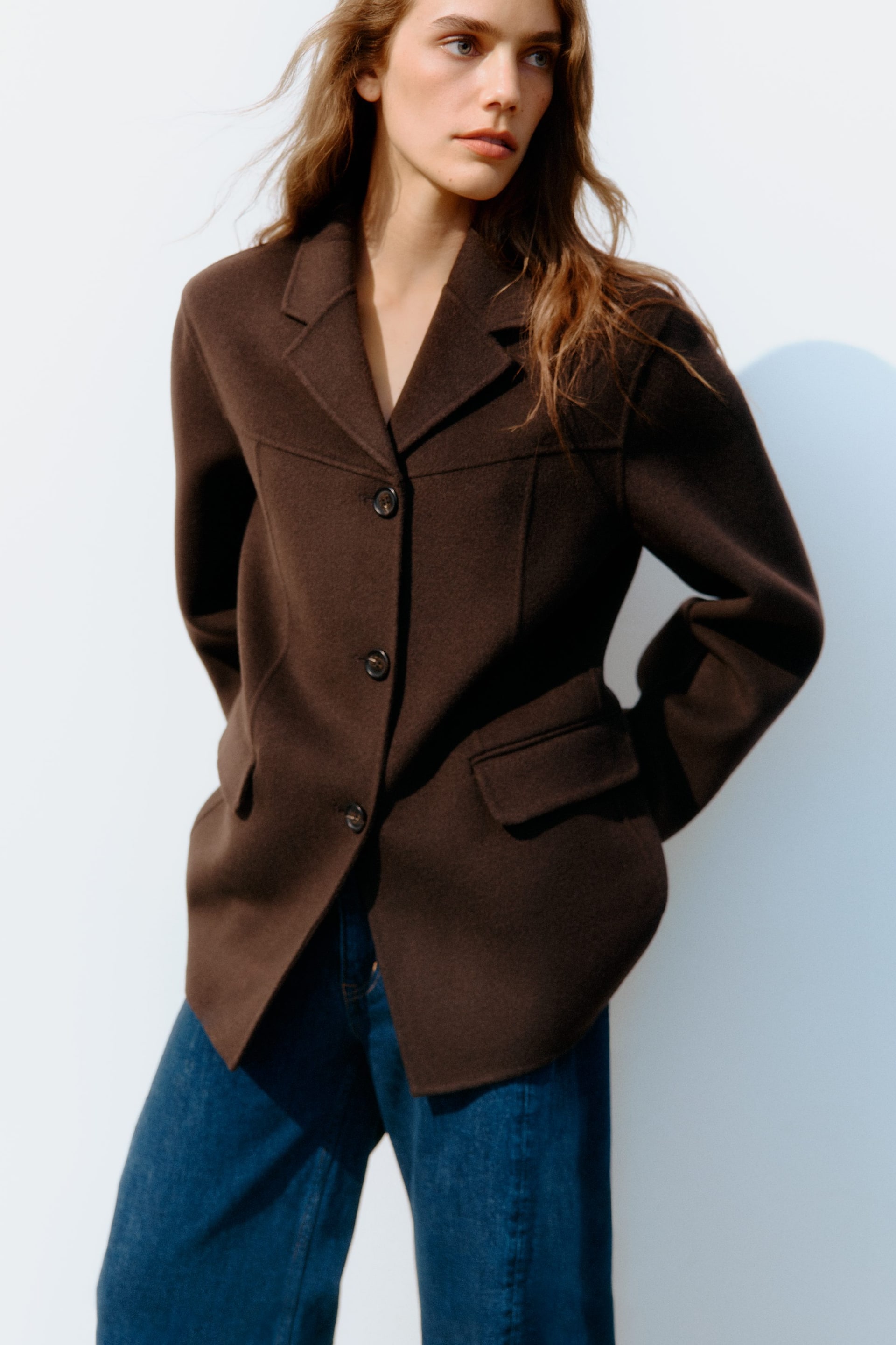 Nagyobb kép megtekintése: Egy sötétbarna, egysoros zakó látható ELÖLRŐL, három sötét gombbal, bevágott hajtókákkal és zsebfedős zsebekkel. A zakó finom szabásvonalakat mutat, és kék, laza szabású farmernadrág fölött van viselve.