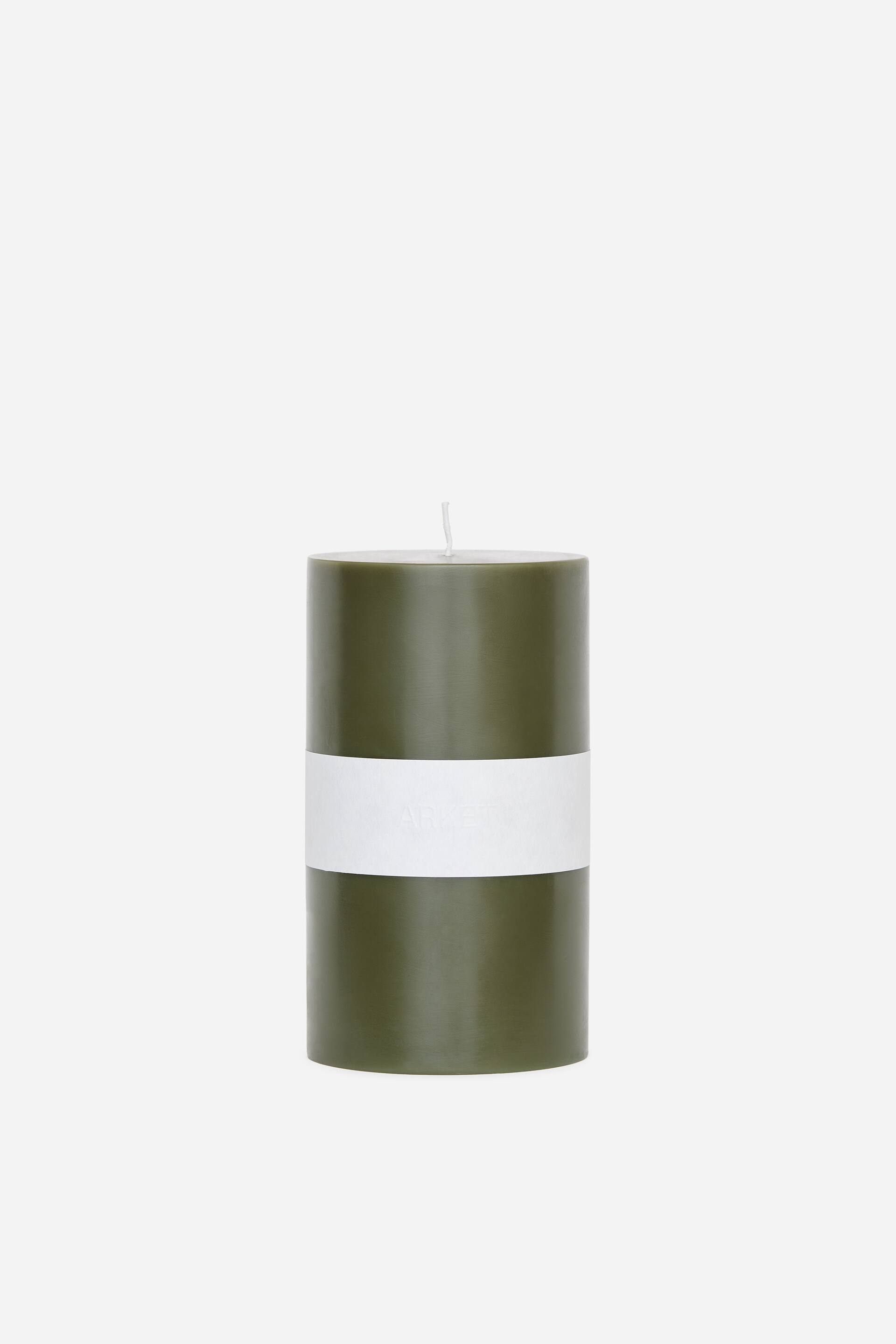 View larger image: A cylindrical olive green wax pillar candle stands upright, featuring a smooth finish and a single wick at the top. A wide white paper band wraps around the centre of the candle.