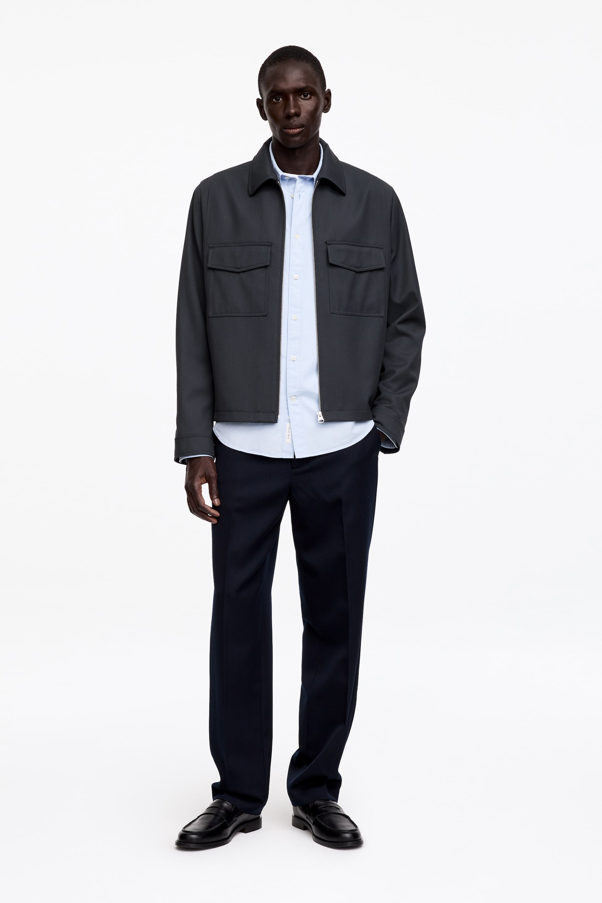 View larger image: A dark grey wool-blend zip jacket, cut for a relaxed fit, is being worn by the model over a light blue shirt. The jacket features a regular collar, a partially unzipped front, and two chest pockets with flaps. The model's hand is resting in a side pocket.