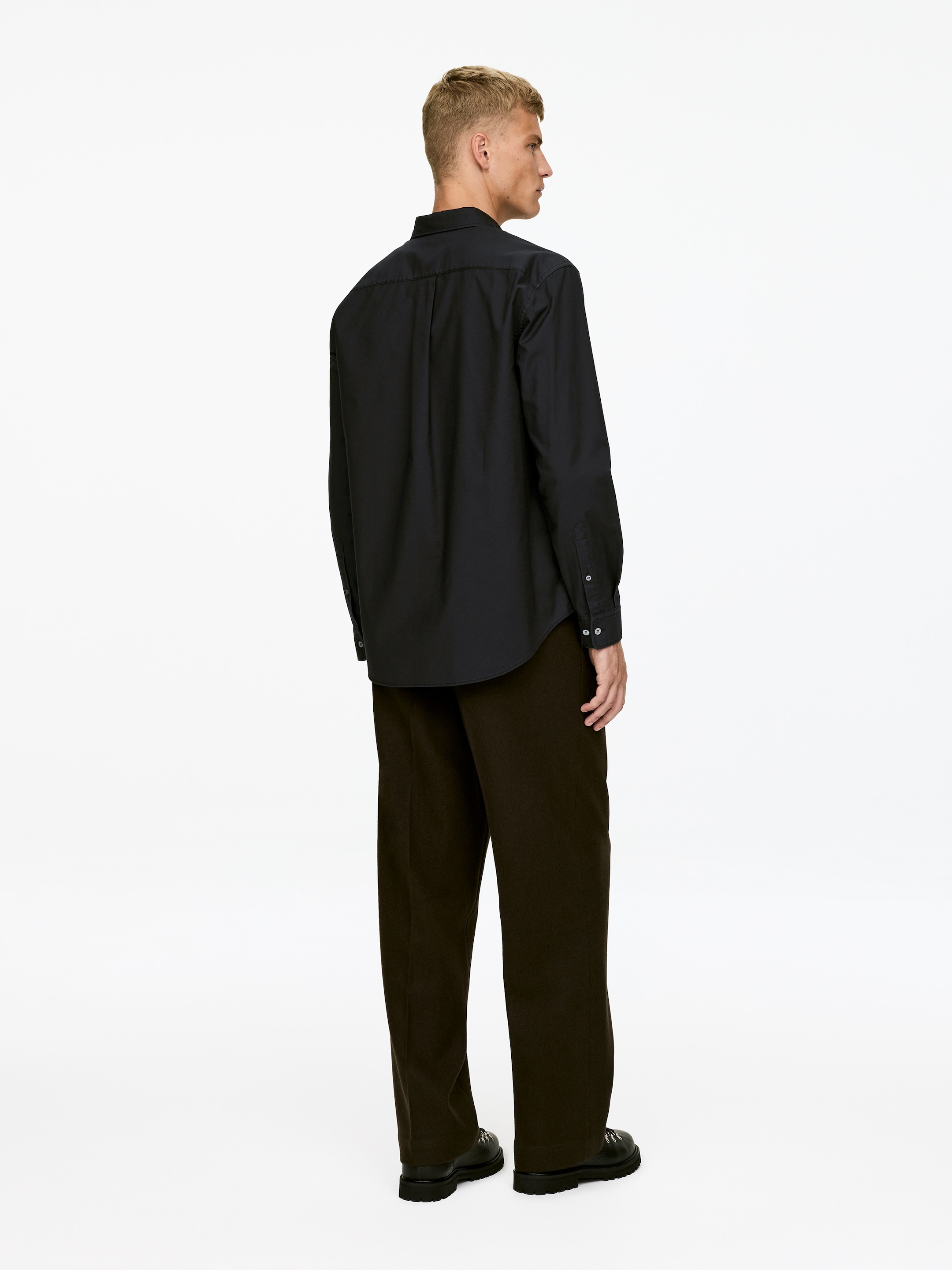 Ver imagen más grande: Una camisa negra de manga larga, vista desde atrás, presenta un canesú trasero, un cuello clásico y puños con botones. Se combina con pantalones marrón oscuro de corte holgado que caen sobre zapatos negros.