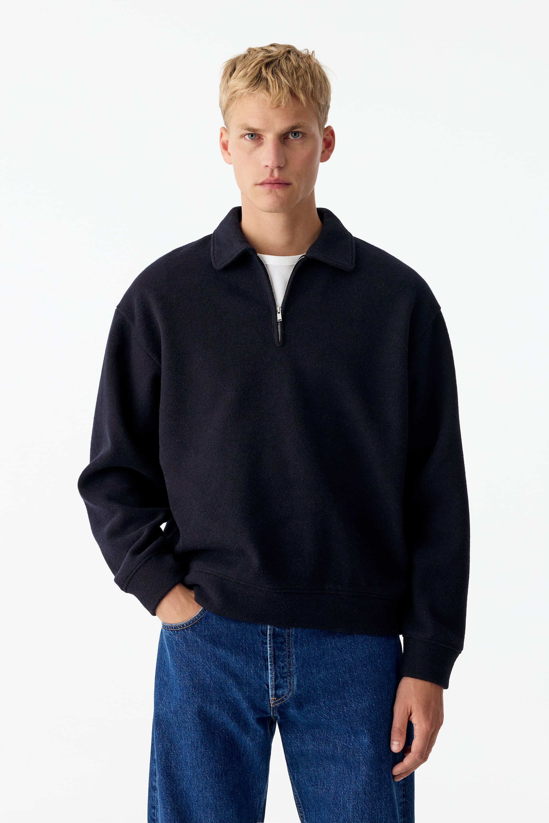 Grotere afbeelding bekijken: Een donker marineblauwe sweatshirt met halve rits, puntkraag en zilverkleurige ritssluiting wordt door het model gedragen over een wit T-shirt met ronde hals. De sweatshirt heeft geribde manchetten en zoom en wordt gecombineerd met een blauwe jeans.