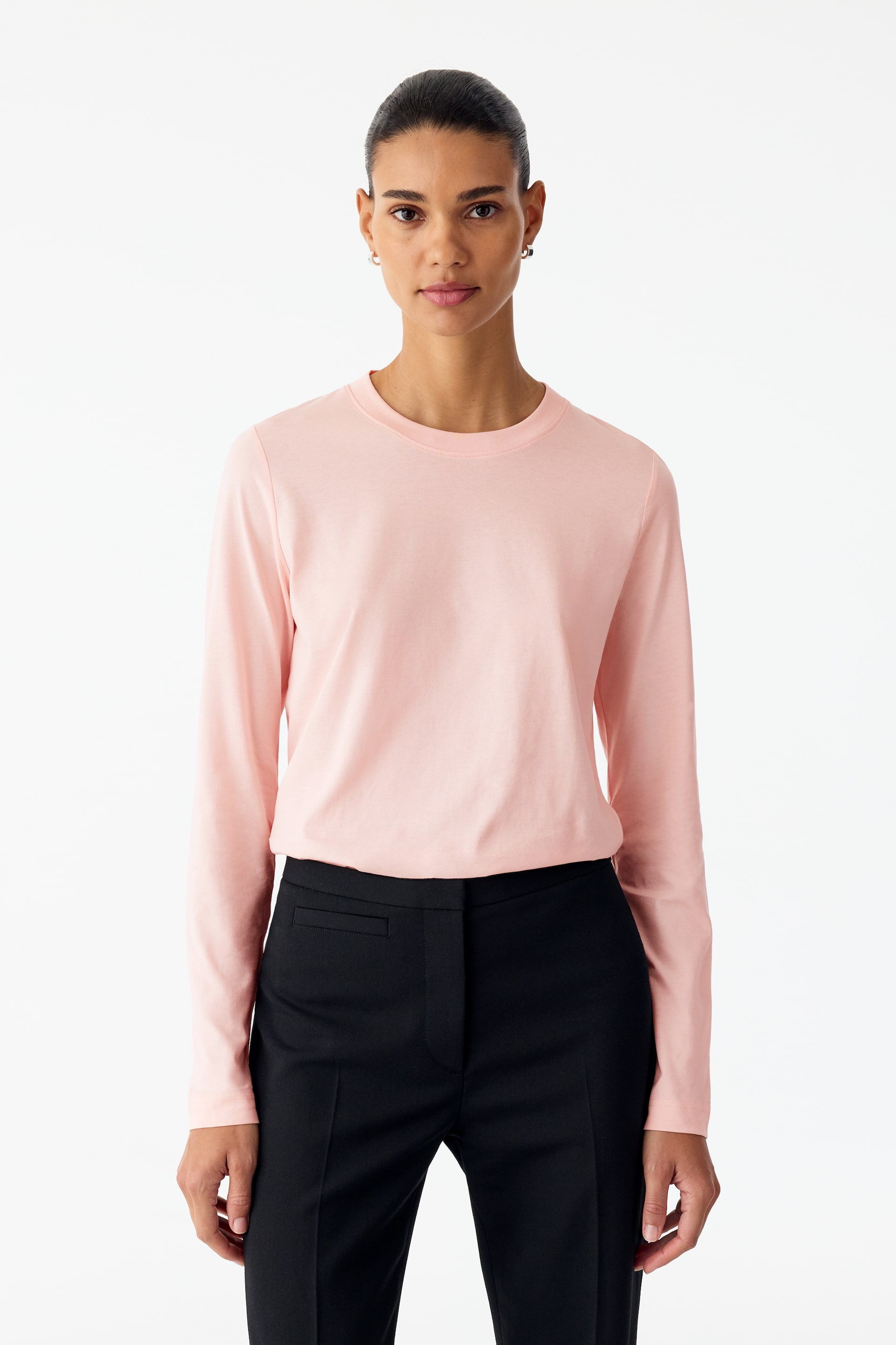 Vezi imaginea mai mare: Un tricou roz pal cu mânecă lungă, cu guler rotund ribat și mâneci fără manșetă, este purtat de model. Cămașa cu croială regular fit este stilizată băgată în pantaloni negri croiți.