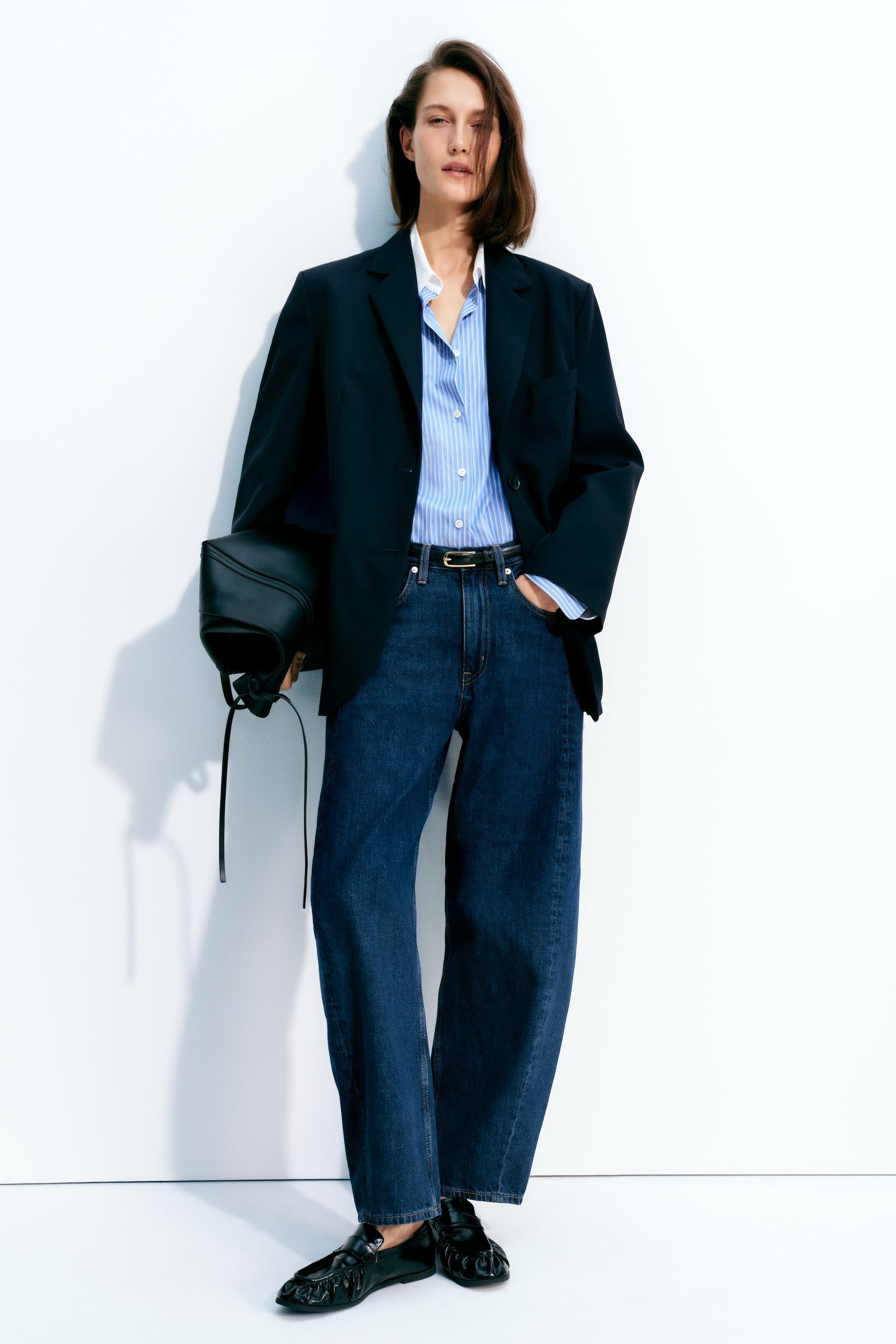 View larger image: An oversized navy blazer is layered over a light blue striped button-down shirt. Dark wash, wide-leg jeans are styled with a thin black belt and black flat loafers featuring a gathered vamp, while a black soft leather bag is held.