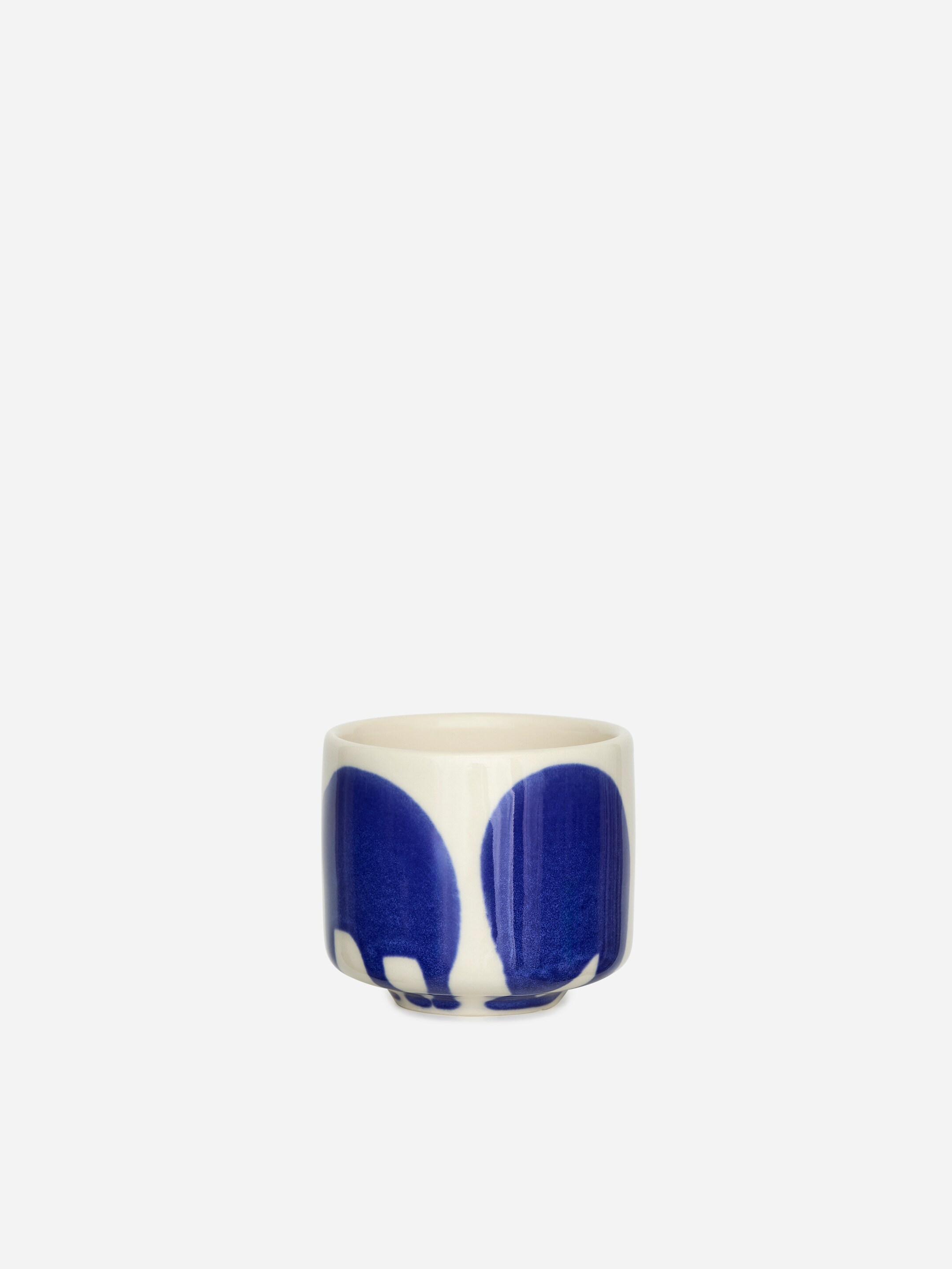 View larger image: A handleless cup is featuring a glazed stoneware finish, with an off-white base and large, irregular blue shapes decorating its exterior, presented FRONT.