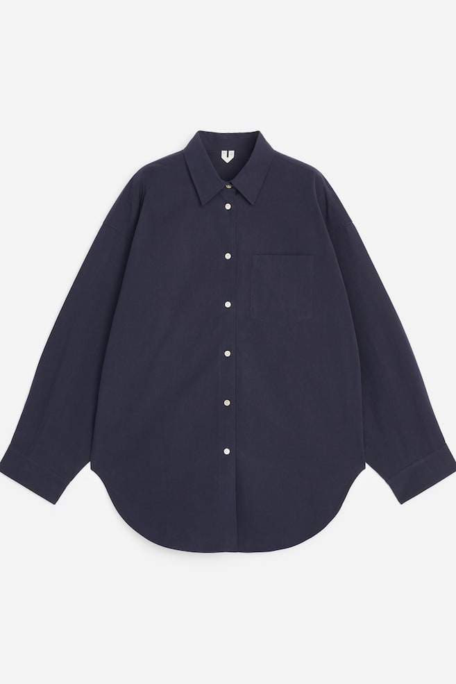 Une chemise bleu foncé surdimensionnée confectionnée en coton pima épais est présentée de face. Elle est dotée de manches en forme de chauve-souris, d'une poche poitrine unique, d'un col pointu et d'une fermeture boutonnée avec des boutons clairs, et se termine par un ourlet incurvé. - 1