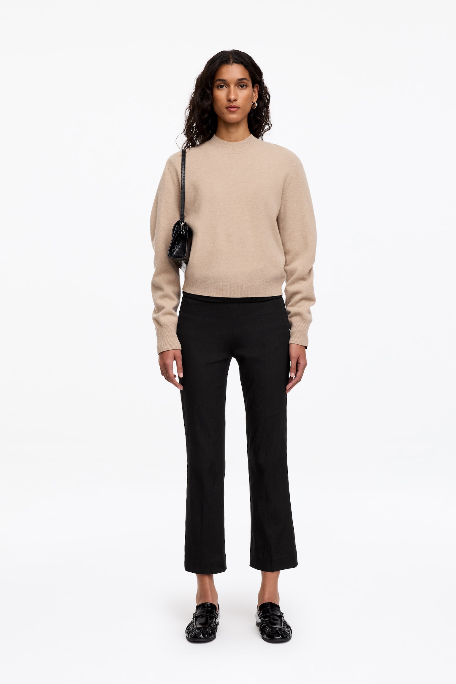 Ce pantalon noir court est porté par le mannequin, avec une silhouette droite et de discrètes plis marqués à lâavant. Il est associé à un pull beige à manches longues et col rond, des mocassins noirs et un sac à bandoulière noir. - 1
