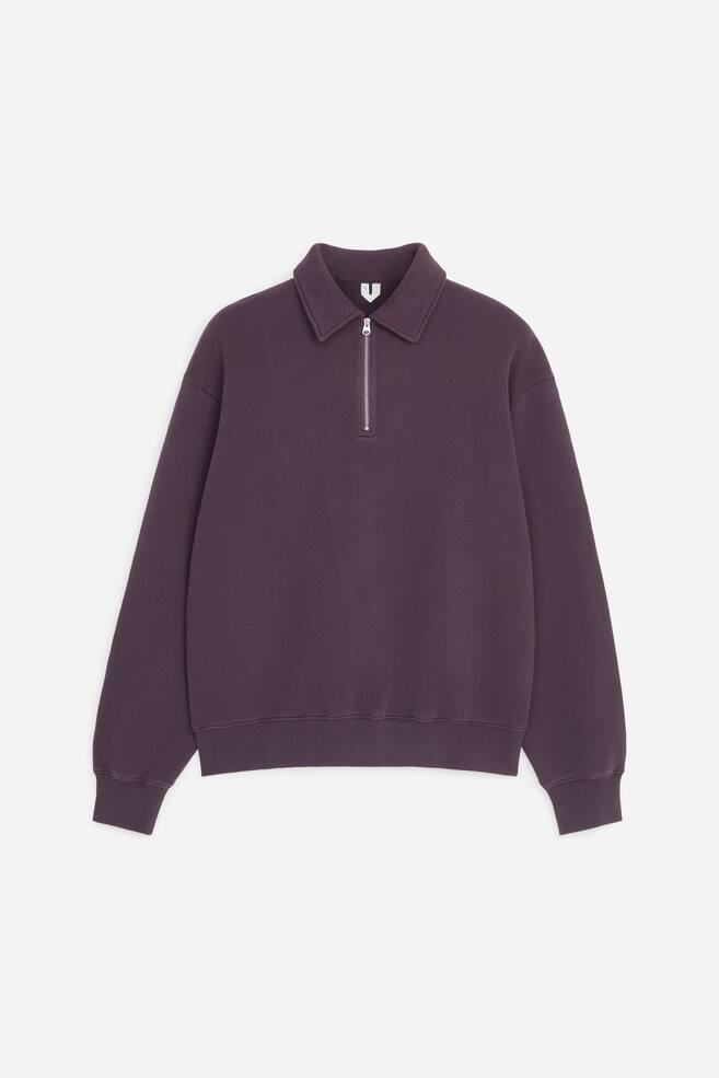 A dark purple sweatshirt is shown front-facing, featuring a classic collar and a silver metal half-zip fastening. The garment has dropped shoulders, long sleeves finished with ribbed cuffs, and a matching ribbed hem. - 1