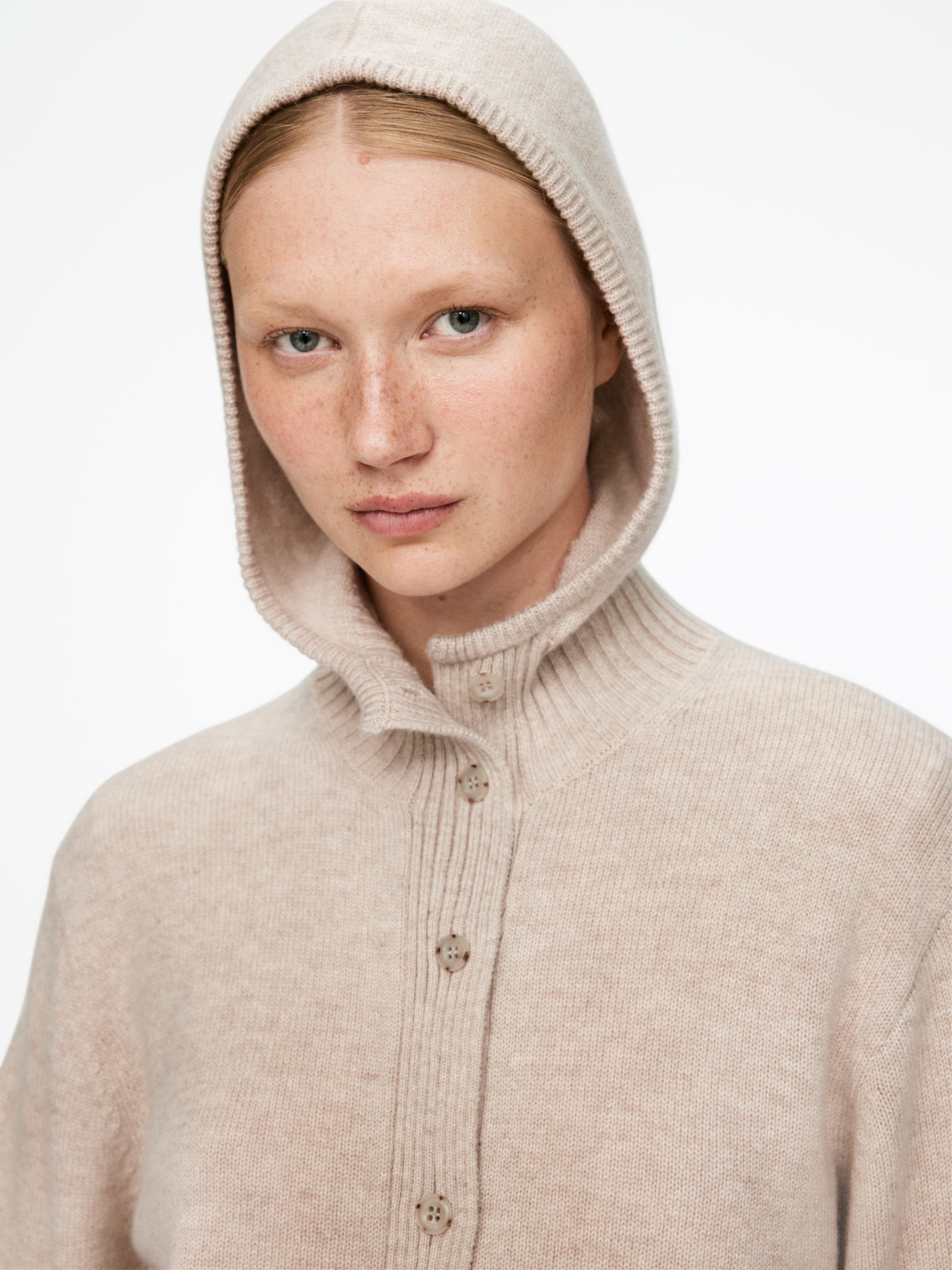 Visualizza immagine più grande: Un cardigan con cappuccio lavorato a maglia beige screziato è indossato, leggermente inclinato verso destra. Presenta un cappuccio integrato tirato su sopra la testa, un colletto alto a coste e un'abbottonatura frontale con diversi bottoni marrone chiaro.