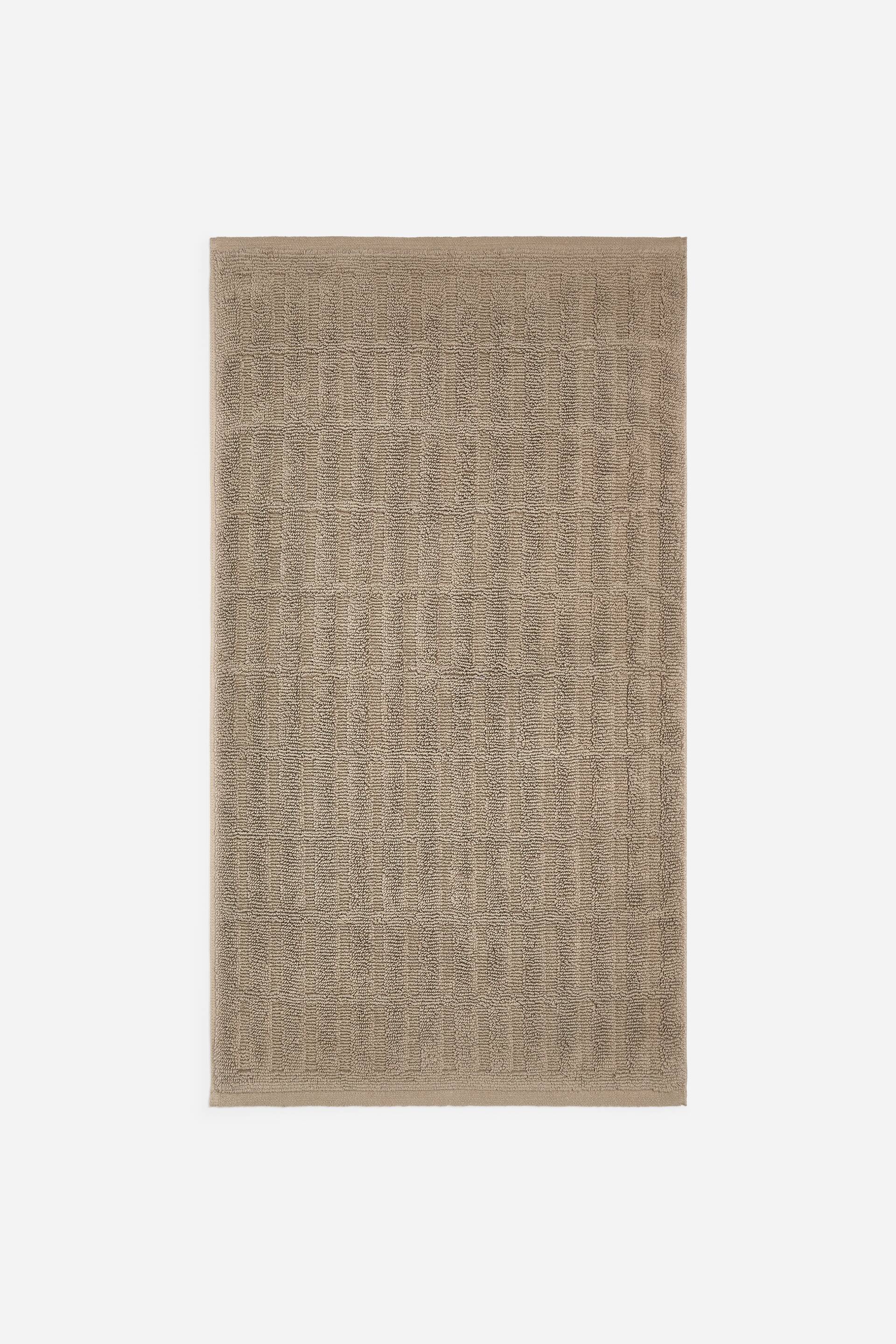 Ver imagen más grande: Una alfombrilla de baño rectangular de rizo de algodón en un tono beige topo aparece extendida en una vista frontal, mostrando un patrón texturizado de cuadrícula en toda su superficie. La alfombrilla presenta bordes de tejido plano en los extremos superior e inferior.