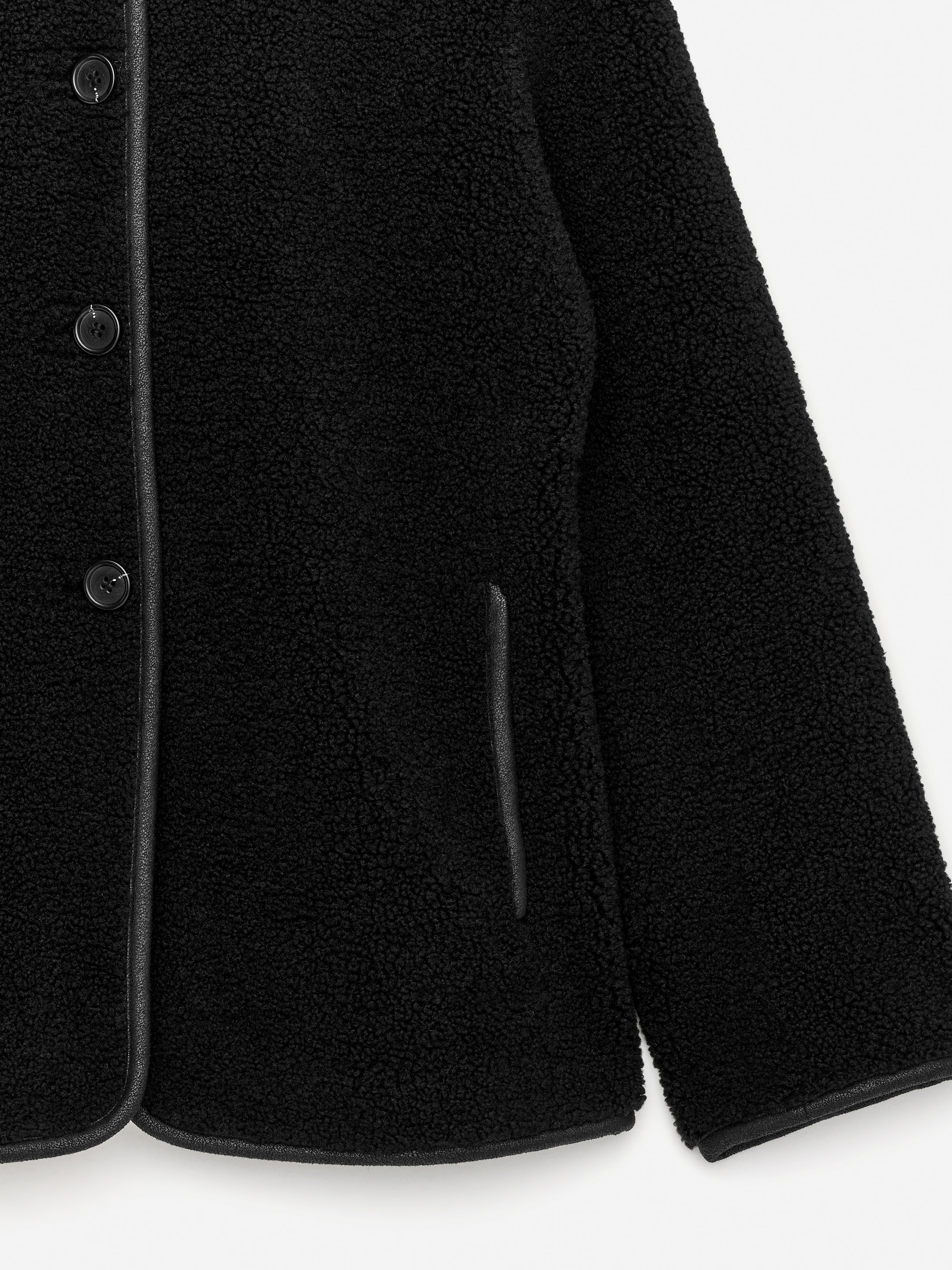 View larger image: A black teddy fleece jacket is featuring black piping along the front button placket, the rounded hem, and a vertical slit pocket opening. Three round black buttons are visible on the placket, and the sleeve cuff is also edged with black piping.