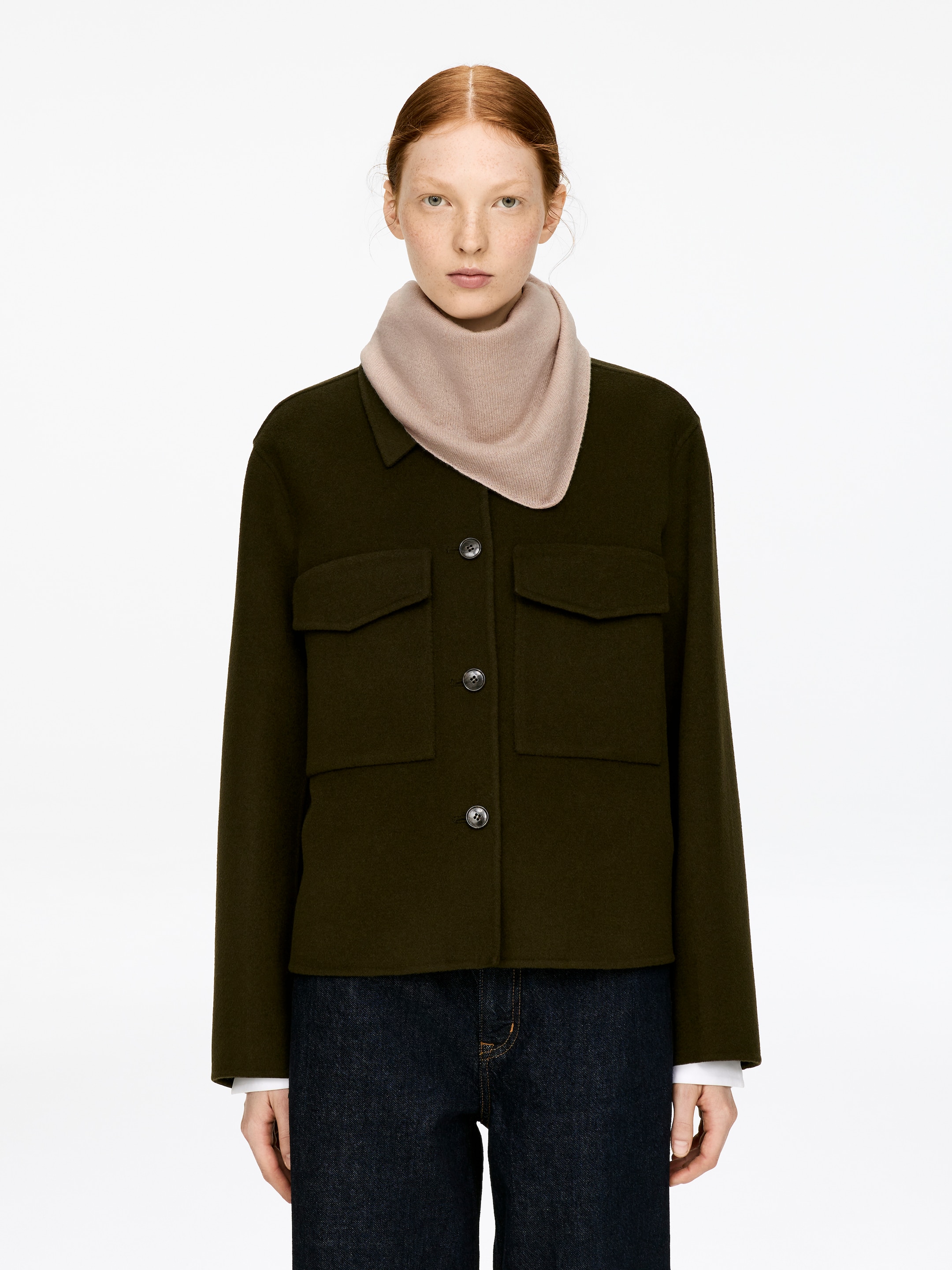 Grösseres Bild ansehen: Kaschmirschal. Ein blassrosa Kaschmirschal wird vom Model getragen, um den Hals in einer dreieckigen Form gestylt, über einer dunkelgrünen Jacke geschichtet.