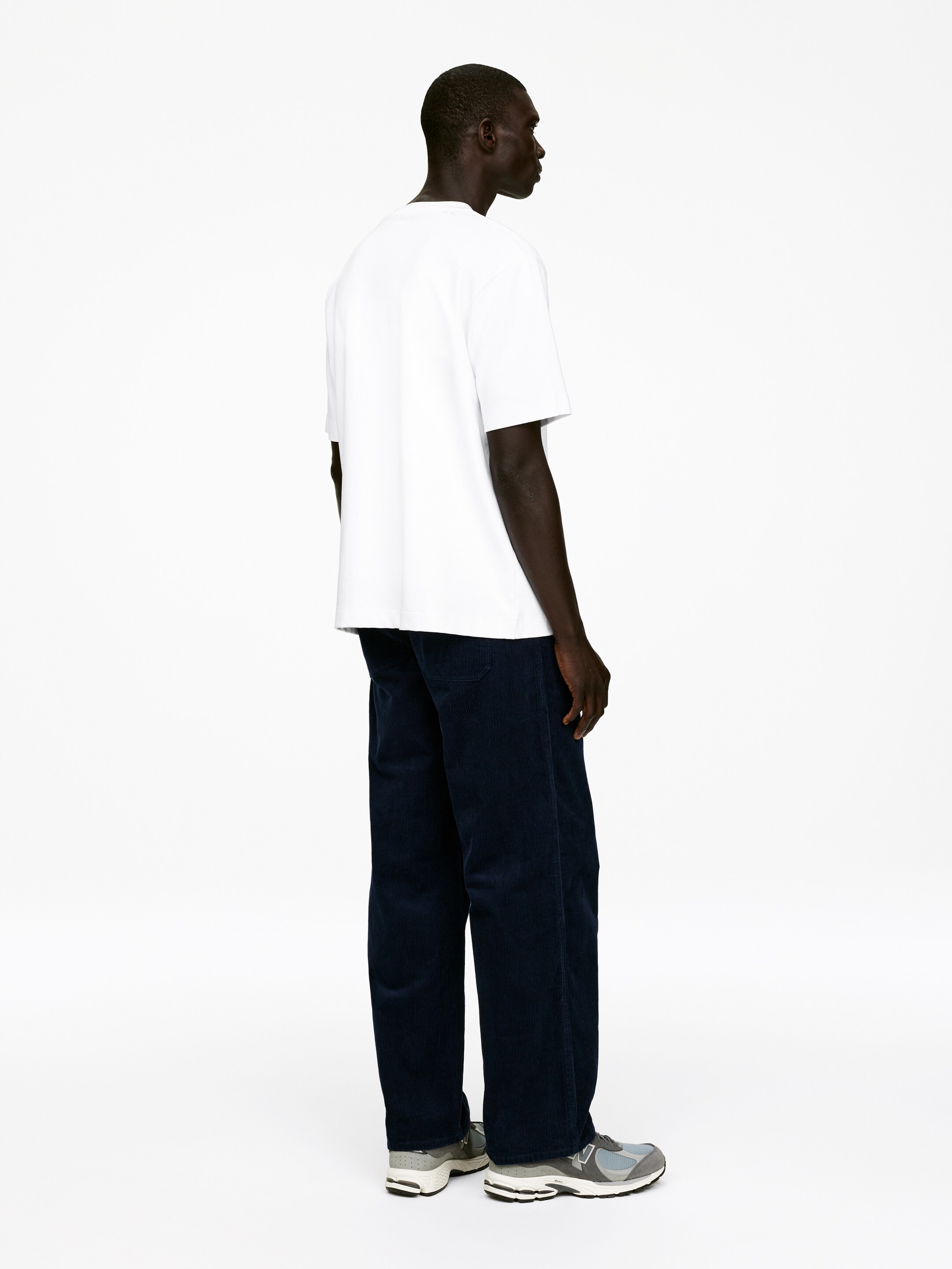 Agrandir l'image: Un t-shirt blanc à col rond et un pantalon en velours côtelé bleu marine à coupe ample sont portés, vus sous un angle arrière. Le t-shirt présente une coupe décontractée, et le pantalon affiche une silhouette à jambes droites avec une texture de velours côtelé visible. Des baskets grises et bleues complètent le look.