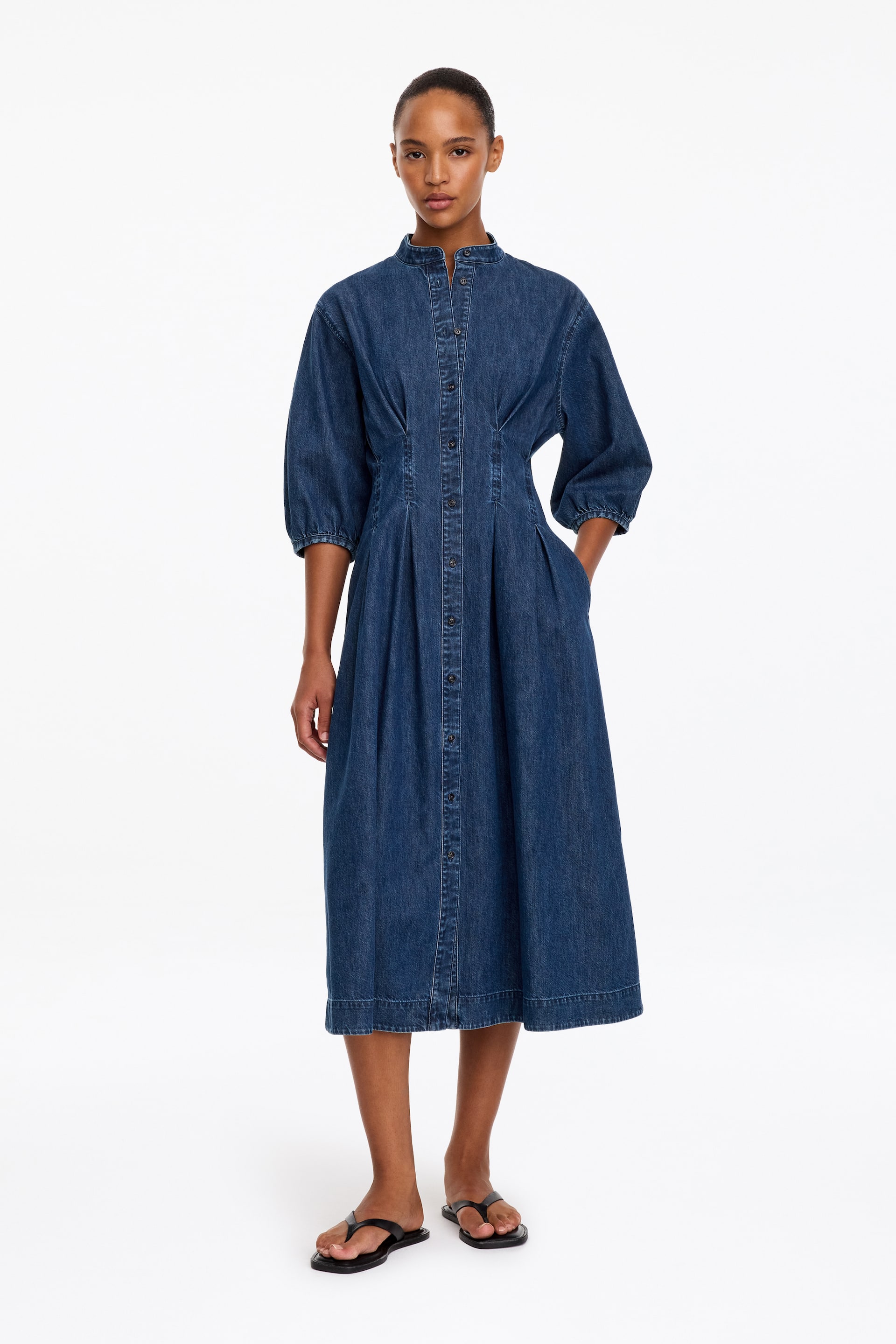 Vis større bilde: En blå denim midikjole bæres av modellen, med oppstående krage, knappelukking foran, rynkede ermer i albuelengde og et plissert liv. Modellen står med den ene hånden plassert i en sidelomme i sømmen.
