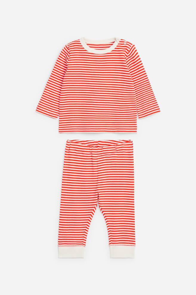 Een tweedelige kinderkledingset is van voren afgebeeld, met een top met lange mouwen en bijpassende broek in een horizontaal rood-wit streeppatroon. De top heeft een crÃ¨mekleurige ronde hals met drukknoopsluiting op de linkerschouder, terwijl de broek is voorzien van een elastische tailleband en crÃ¨mekleurige geribde enkelboorden. - 1