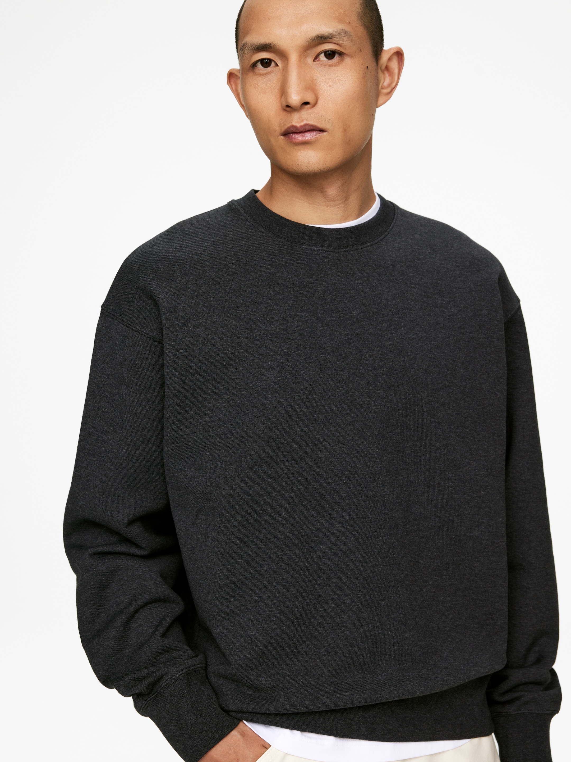 Größeres Bild ansehen: Ein meliertes dunkelgraues Sweatshirt mit Rundhalsausschnitt wird getragen, nach vorne gerichtet, mit einer entspannten Passform, gerippten Bündchen und einem gerippten Saum. Ein weißes Unterhemd schaut aus dem Kragen und unter dem Saum hervor.