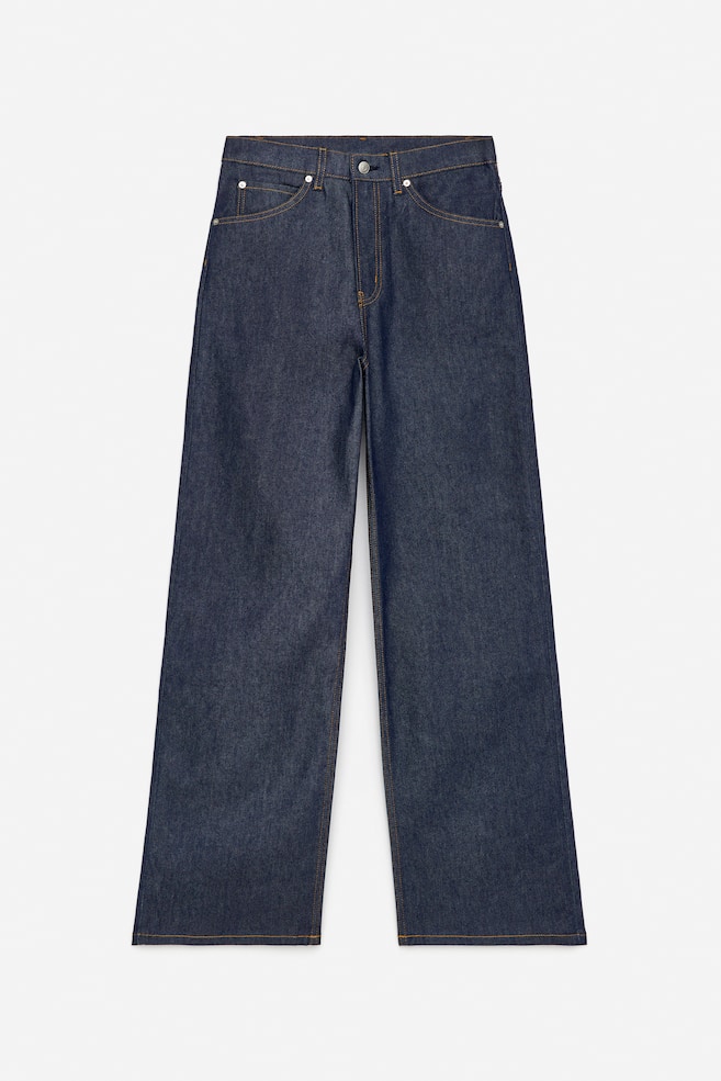 Een paar donkerblauwe denim jeans wordt van de VOORKANT gepresenteerd, met een hoge taille, een ritssluiting en voorzakken met klinknagels. De jeans is ontworpen met een slanke pasvorm die uitloopt in wijde pijpen, en ze zijn gedetailleerd met zichtbare contrasterende stiksels. - 1