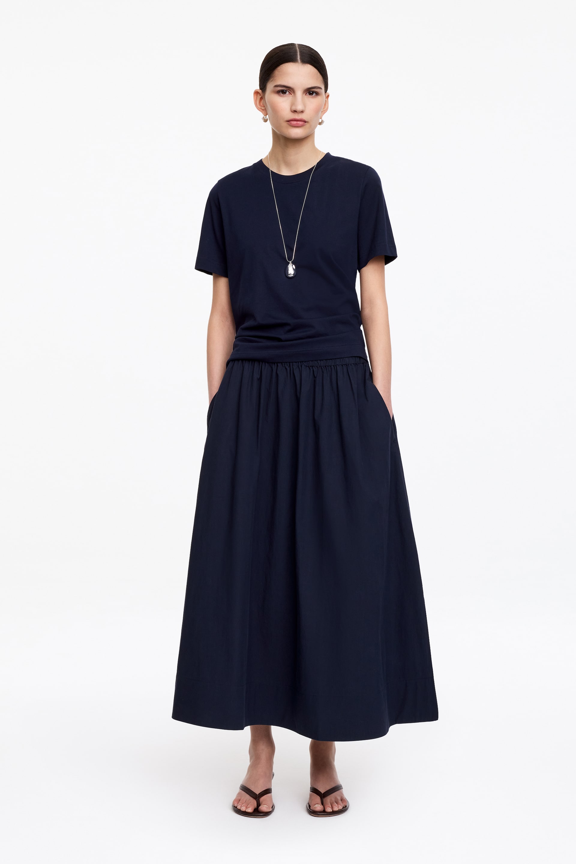 Vis større bilde: En marineblå kortermet t-skjorte bæres sammen med et matchende marineblått skjørt i full lengde, med samlet midje og sidelommer, presentert fra en frontvisning. Brune flate tåsandaler kompletterer antrekket, accessorert med et langt sølvfarget anhengsmykke og små hoop-øreringer.