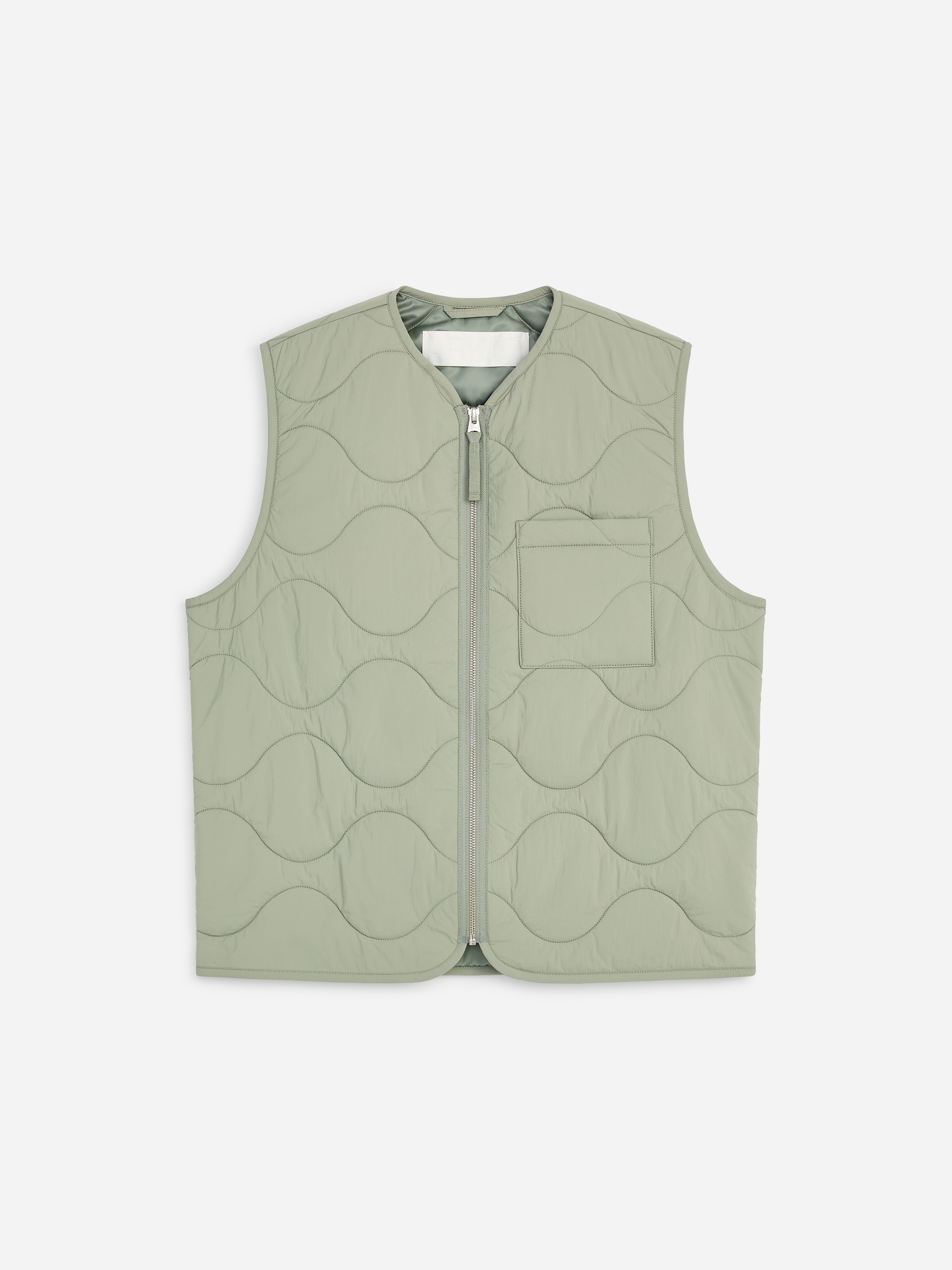 Un gilet matelassÃ© vert clair, ornÃ© dâun motif matelassÃ© en oignon et dotÃ© dâune seule poche poitrine, est portÃ© par le mannequin. Le gilet est portÃ© ouvert par-dessus un sweat-shirt gris clair Ã  demi-zip et associÃ© Ã  un pantalon blanc cassÃ©.