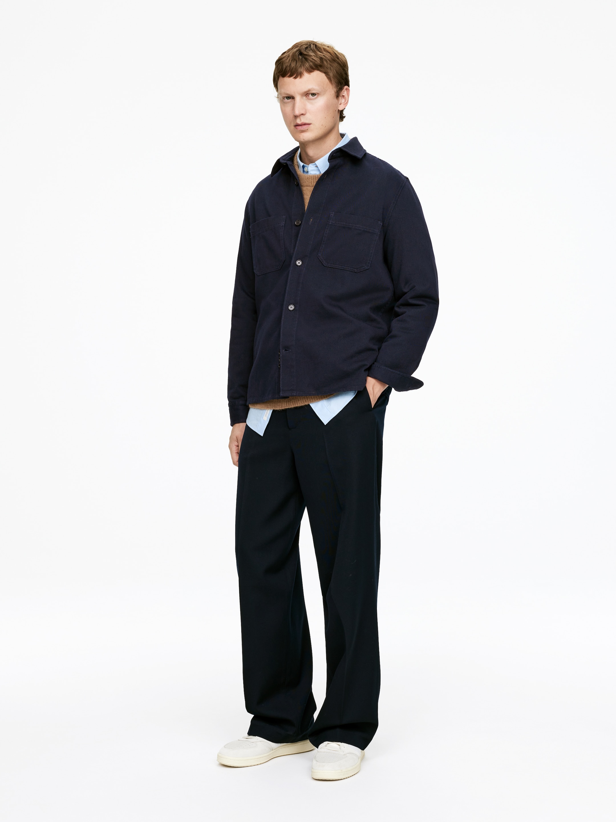 Se større billede: En mørkeblå twill overshirt med almindelig krave, to brystlommer og knappede manchetter bæres af modellen, stylet åben over en lyseblå skjorte og en brun sweater.