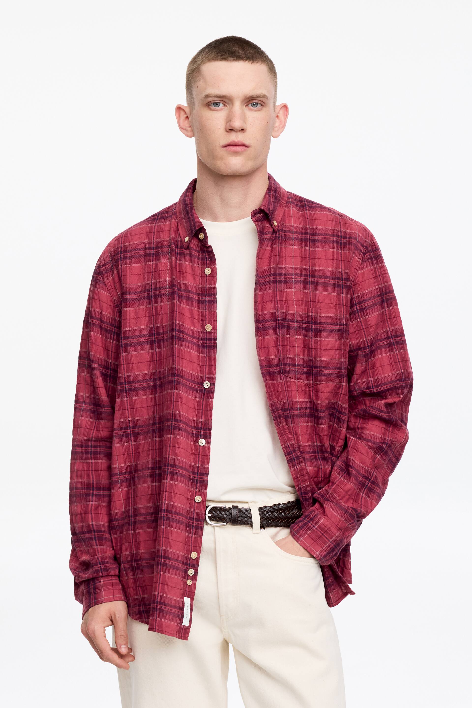 Größeres Bild ansehen: Ein rot und dunkelrot kariertes Button-Down-Hemd wird präsentiert, das über einen Button-Down-Kragen, lange Ärmel mit Knopfmanschetten und eine Frontknopfleiste mit hellen Knöpfen verfügt. Es wird offen über einem weißen T-Shirt getragen und zeigt zwei Brusttaschen.
