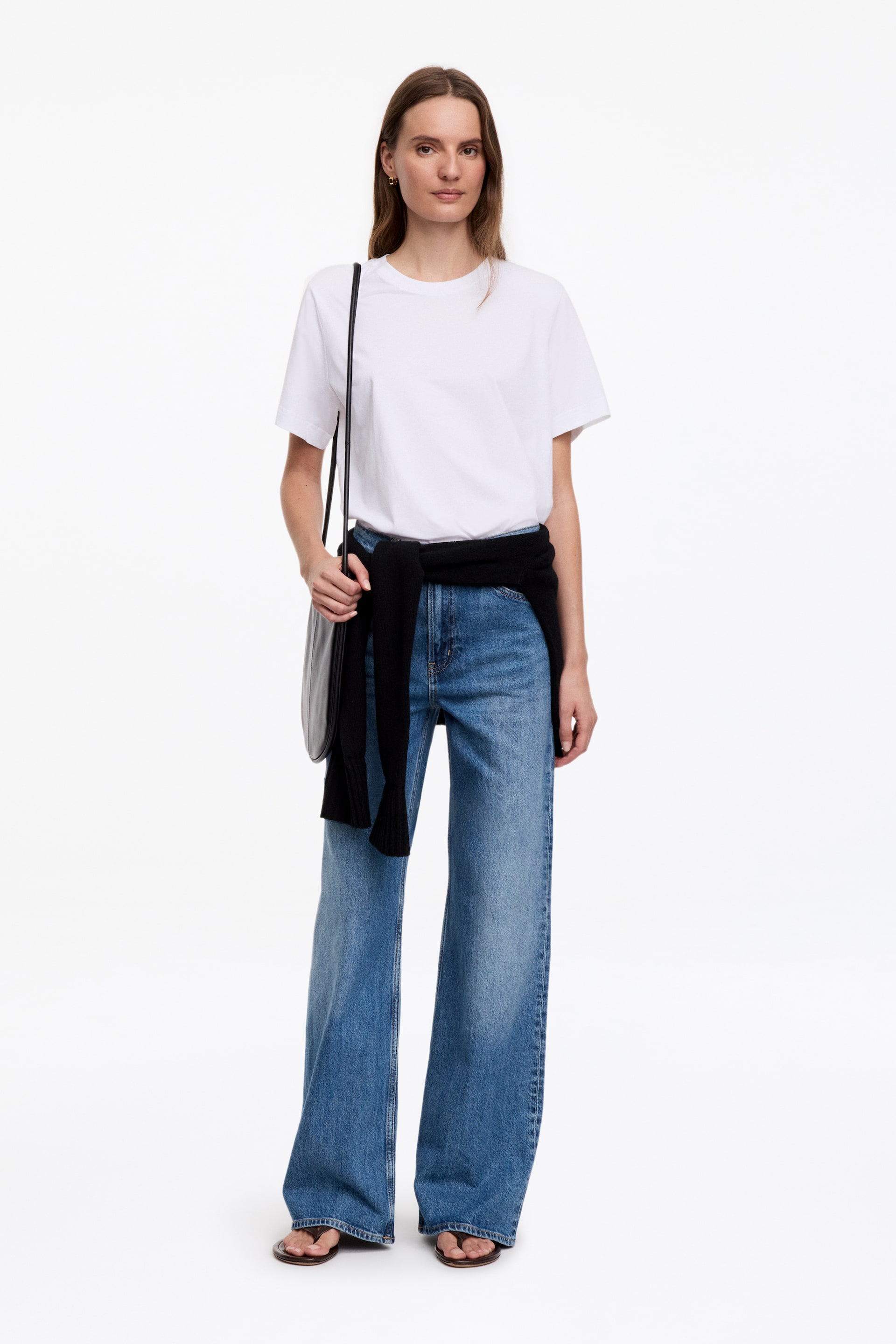 Größeres Bild anzeigen: LUPINE-Jeans sind blaue Denim-Jeans mit hoher Taille, einem schmalen Schnitt an den Oberschenkeln und ausgestellten Beinen. Das Model trägt die Jeans, die mit einem um die Taille gebundenen schwarzen Pullover gestylt sind.