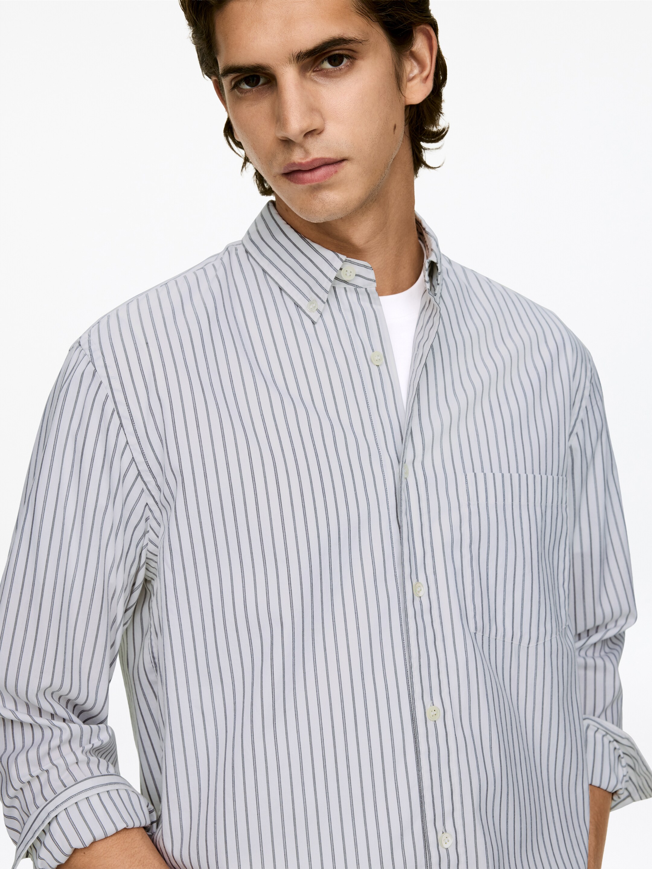 Visualizza immagine più grande: Una camicia bianca a righe verticali è presentata frontalmente con un leggero angolo, con sottili strisce verticali scure, un colletto button-down e una tasca a toppa sul petto a sinistra. Le sue maniche lunghe sono arrotolate e la camicia è indossata sopra una t-shirt bianca con scollo rotondo.