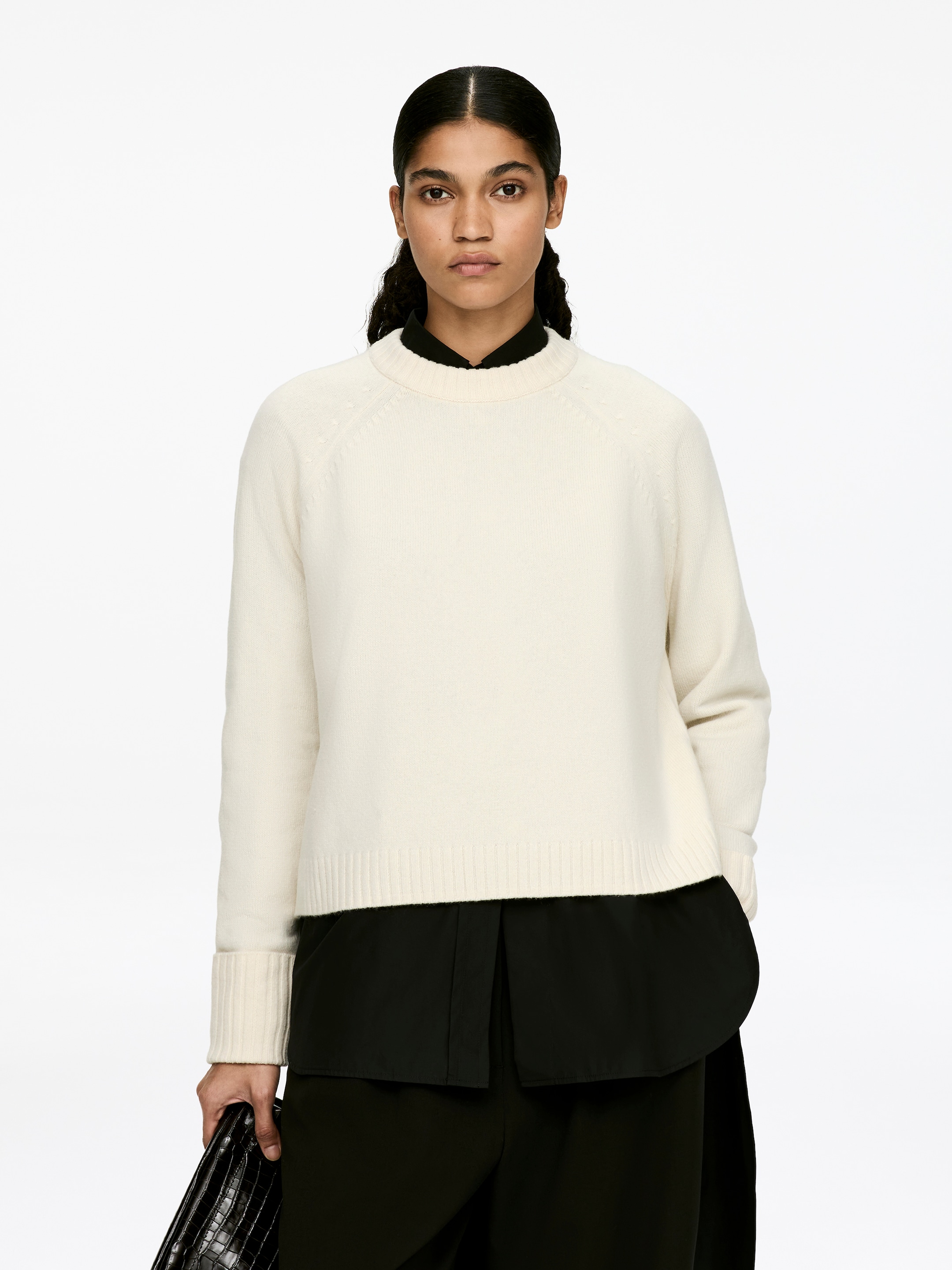 Agrandir l'image: Un pull en laine de couleur crème avec un col rond, doté de manches raglan et de poignets pliés côtelés, est porté par le mannequin. Il est superposé à une chemise noire à col, qui est visible au niveau de l'encolure et de l'ourlet.