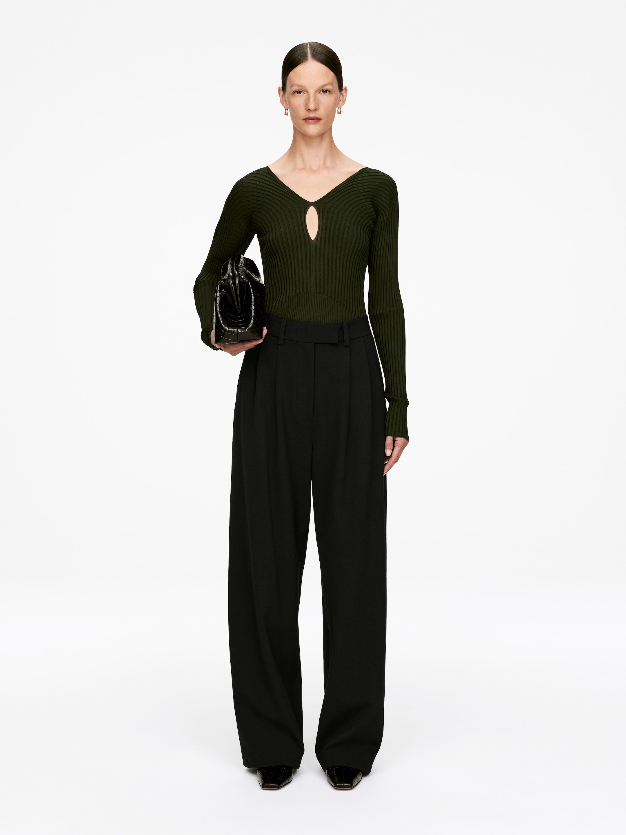 View larger image: A dark olive green ribbed knit top, featuring a V-neckline with a keyhole cutout and long sleeves, is being worn with black high-waisted, wide-leg trousers with front pleats. A black textured handbag is being held, and black square-toed shoes are visible.
